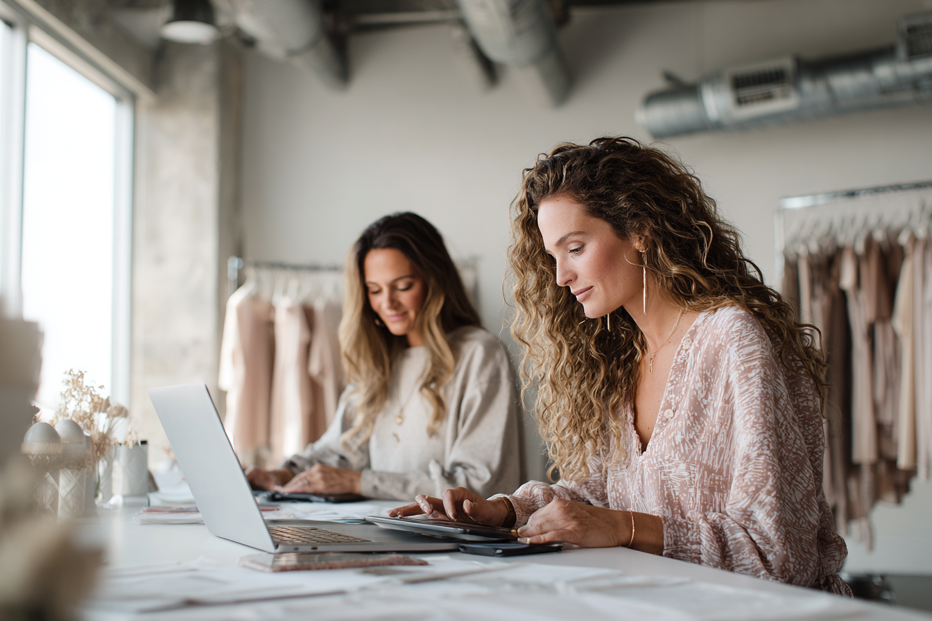 Two women sit at a table working on laptops in a well-lit room with clothing racks in the background.