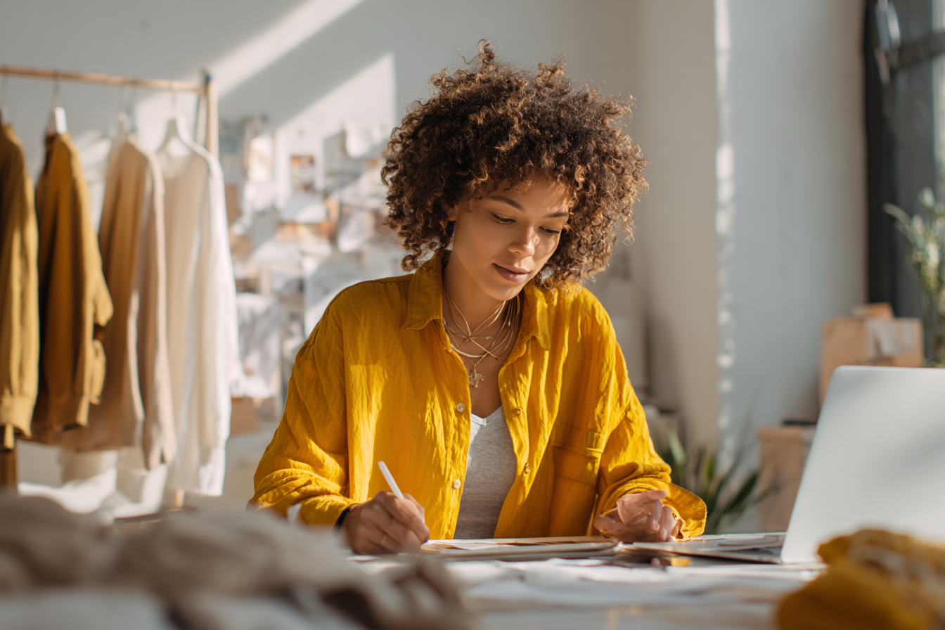 A woman with curly hair wearing a yellow shirt is working at a desk with a notebook, pen, and laptop in a well-lit studio or workshop space.