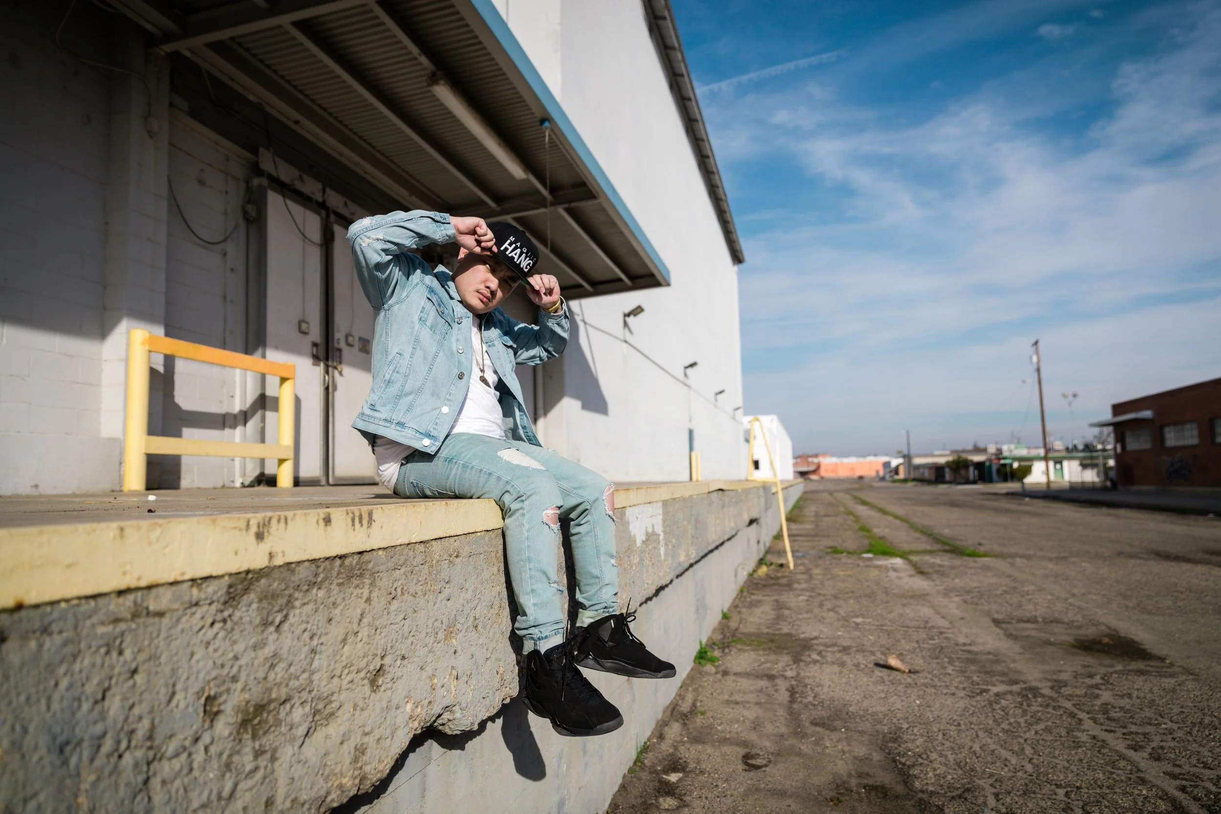 A young person sitting on a ledge outside an industrial building with a cloudy blue sky in the background.
