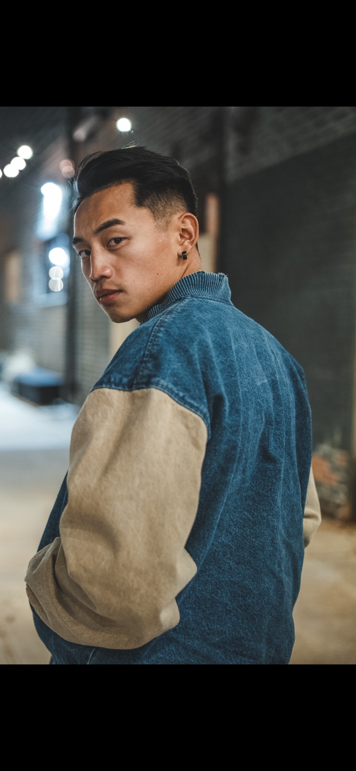 Young man with black hair and an earring, wearing a denim and beige jacket, standing in an urban alley at night.