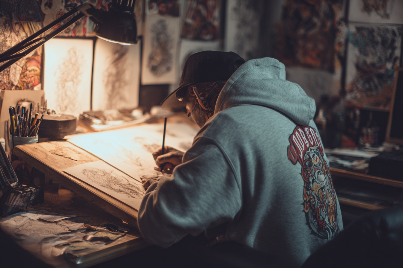 An artist working on a detailed drawing at a cluttered desk in a dimly lit studio, with artwork and sketches on the walls and shelves in the background.