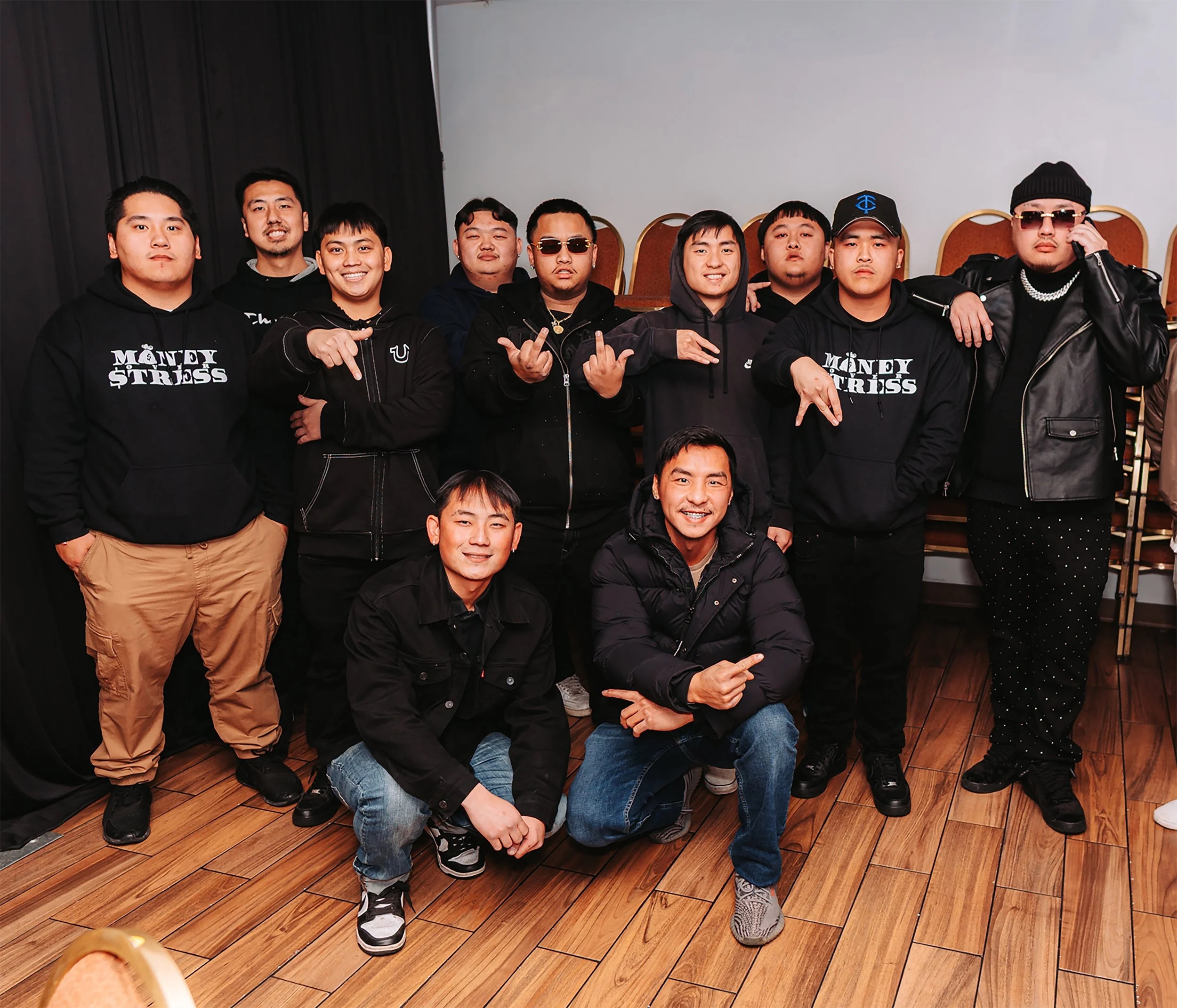 A group of twelve young men posing together indoors, some making hand signs, all dressed in casual streetwear, with a wooden floor and chairs stacked in the background.