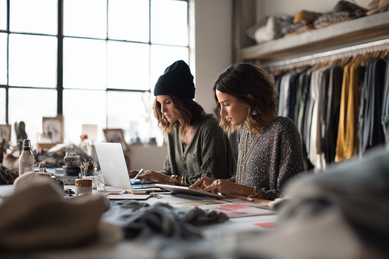 Two women working at a table filled with fabric and design supplies in a bright, industrial-style studio.