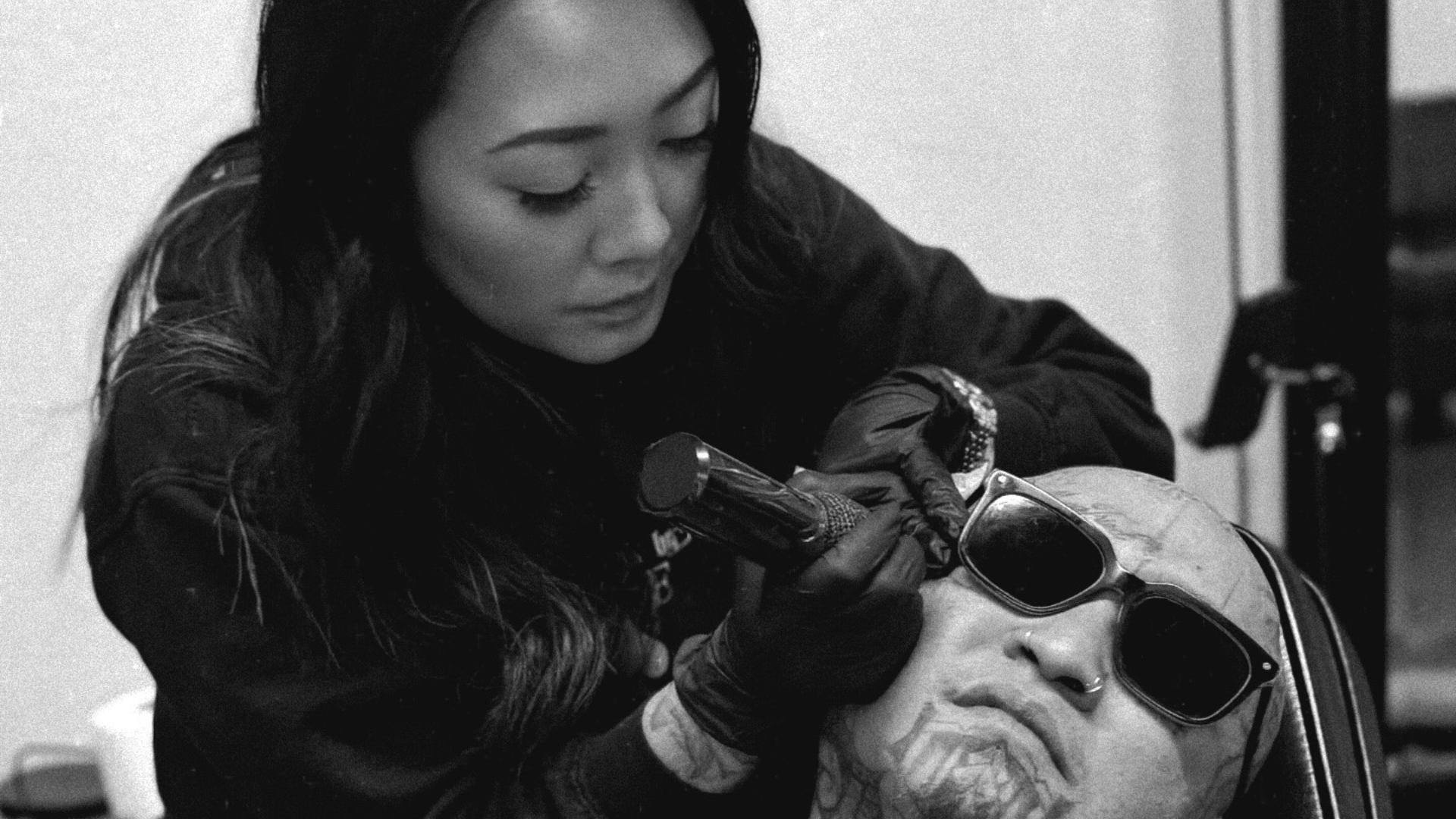 A woman with long dark hair and gloves is tattooing a face sculpture with sunglasses on a chair.