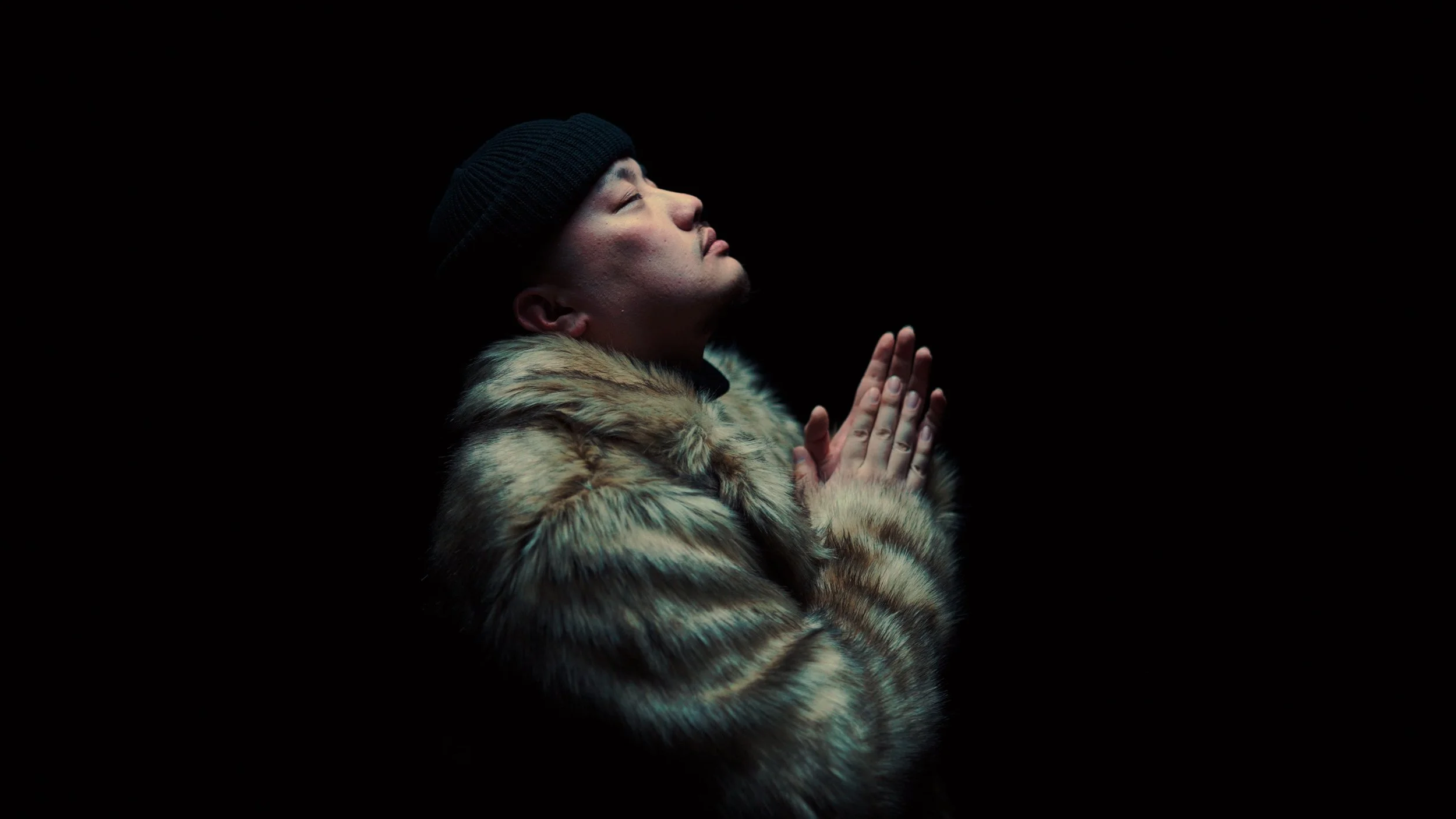 A man in a black beanie and fur coat with his hands together in prayer against a dark background.