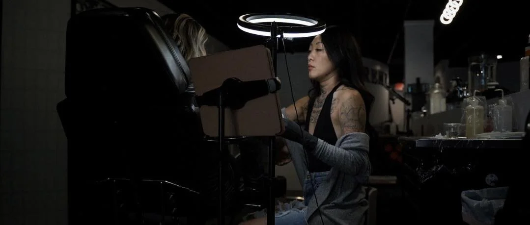A woman with tattoos and black hair sitting at a bar or work station, focused on a task, with a ring-shaped light above her, surrounded by bottles and equipment.