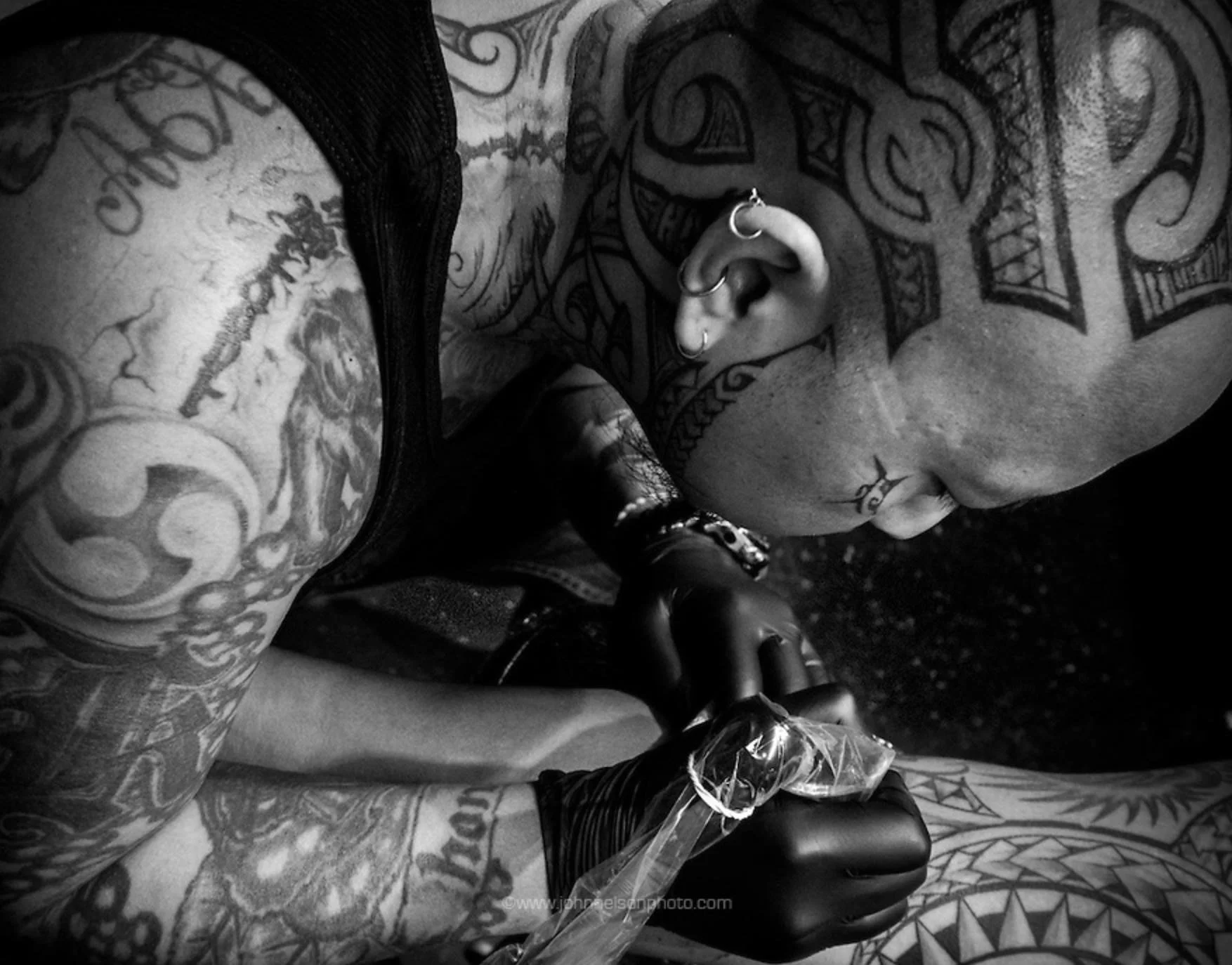 Close-up of a person with numerous tattoos and piercings, wearing a black glove, holding a tattoo machine, with their head tilted downward, surrounded by a dark, tattooed environment.
