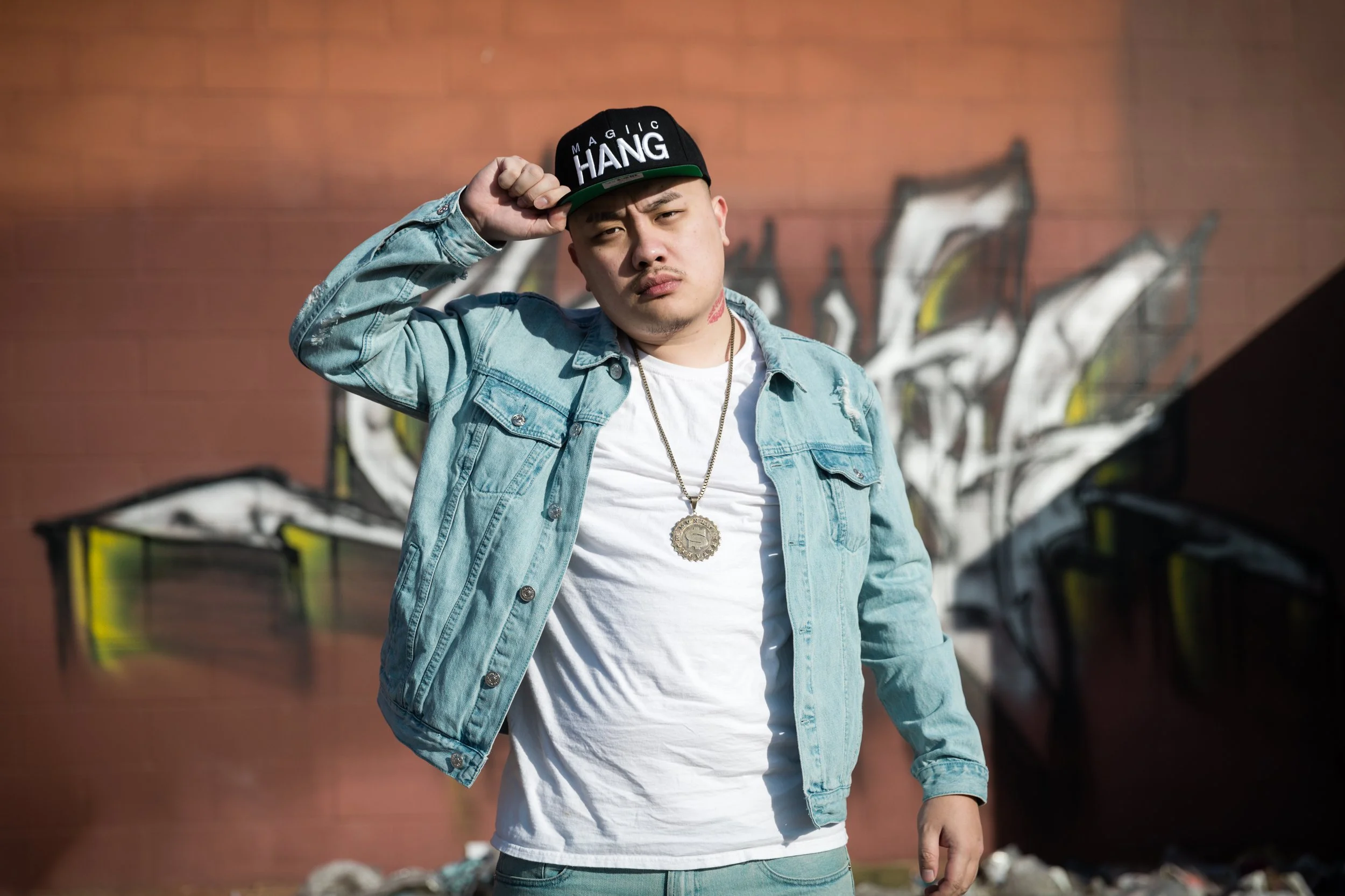 Young man in a denim jacket, white T-shirt, and cap, standing in front of a graffiti-covered brick wall, adjusting his cap.