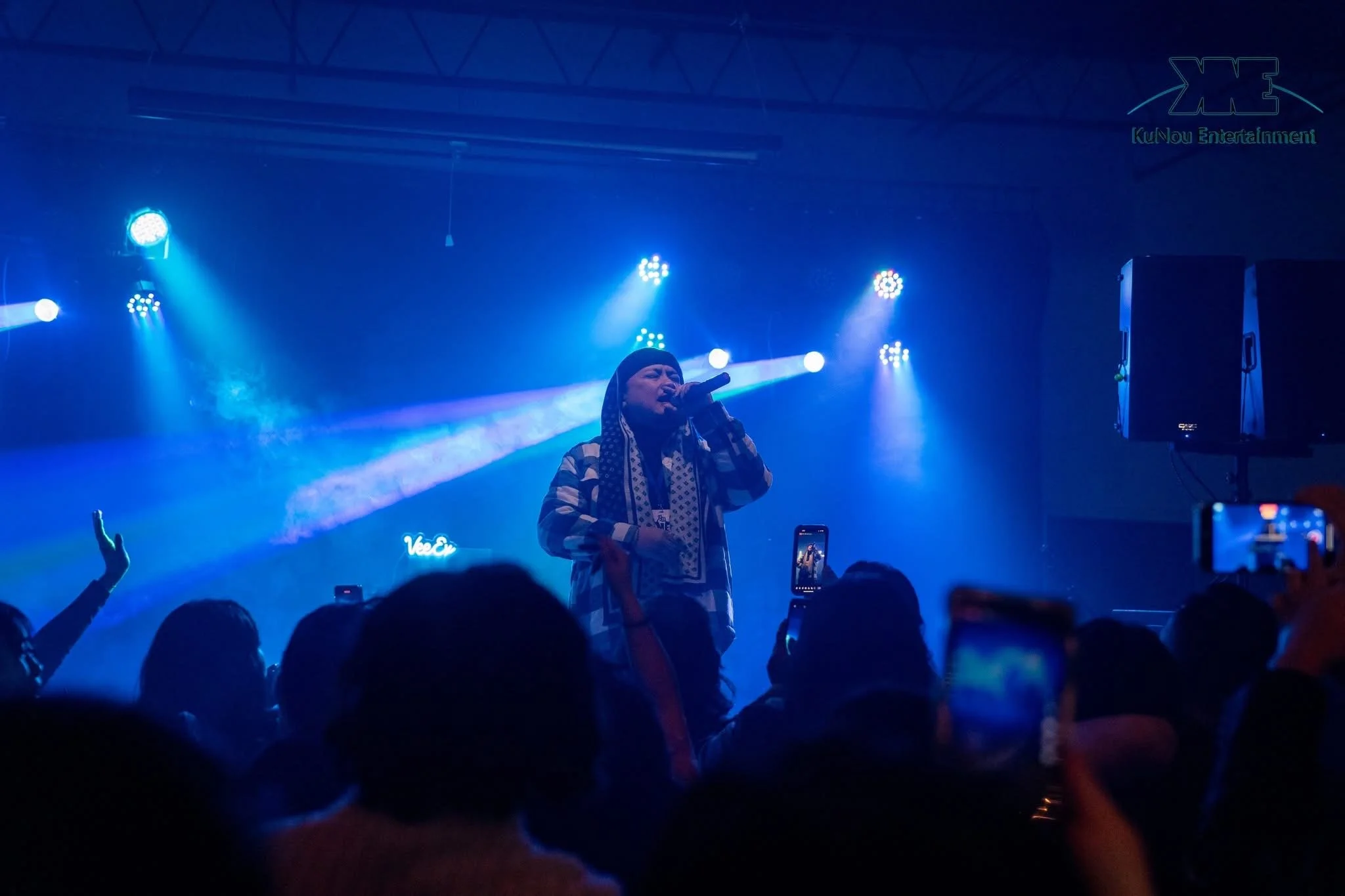 A performer singing into a microphone on stage during a concert, with blue stage lighting and audience members taking photos or videos.