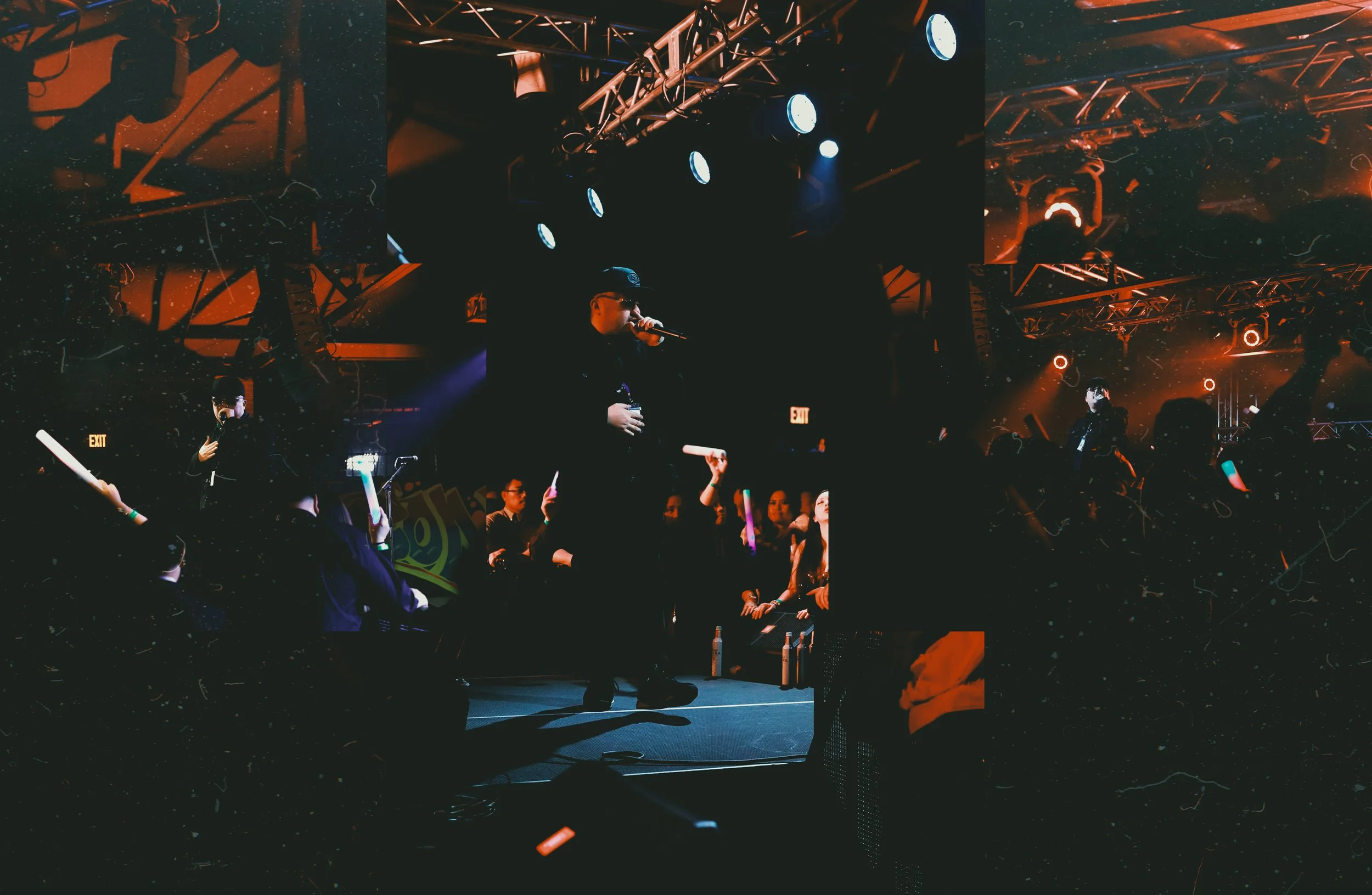 A performer on stage at a concert, wearing a black outfit and cap, singing into a microphone under blue stage lights, with audience members nearby. The venue has a dark, atmospheric ambiance with stage lighting and equipment visible.