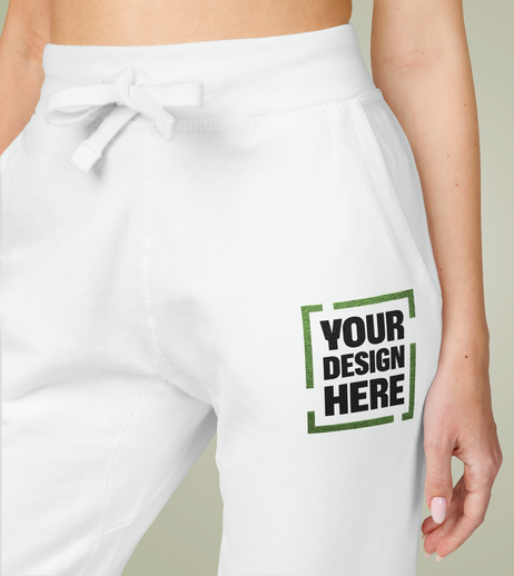 Close-up of a person wearing white sweatpants with a customizable design area and a drawstring waistband. The person's hand is partially visible, resting beside the pants.