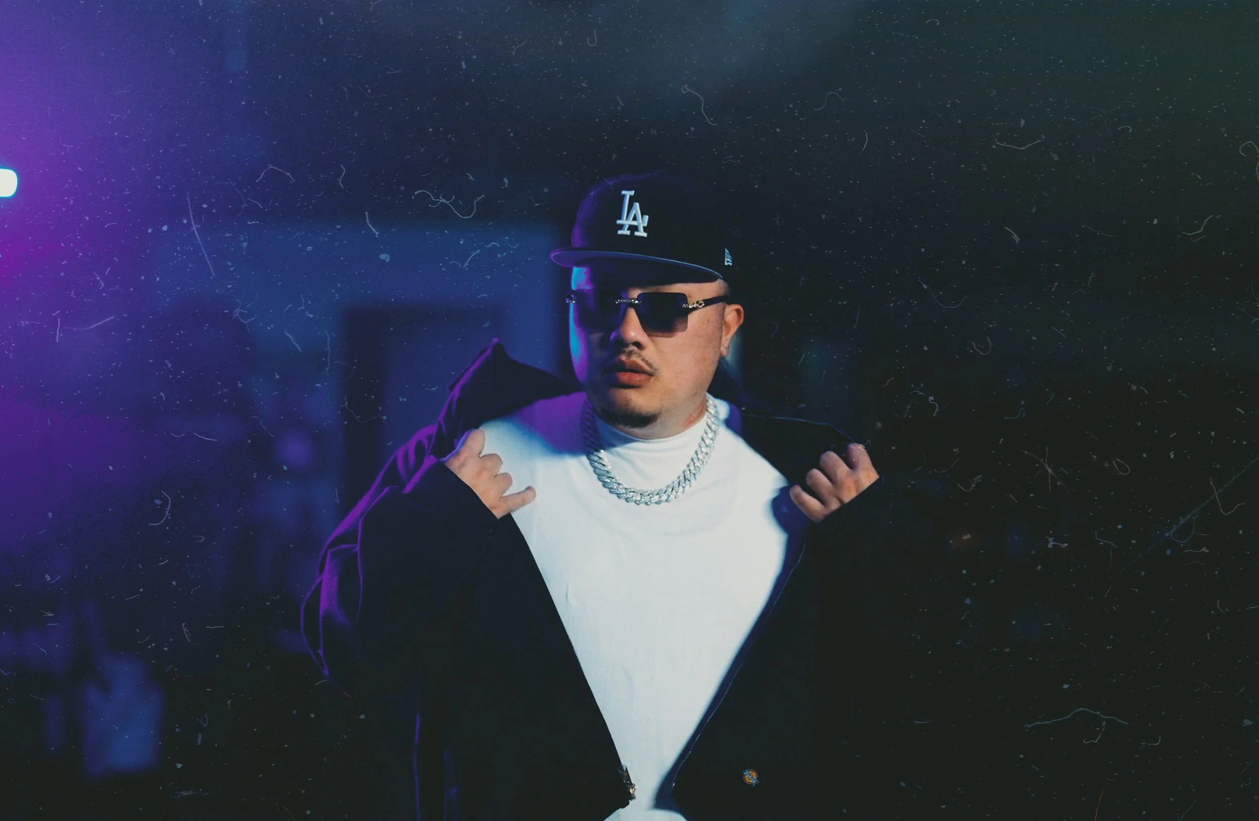 A man wearing a black Los Angeles Dodgers cap, sunglasses, a white shirt, a silver chain necklace, and a black jacket. He is holding the collar of his jacket with both hands.