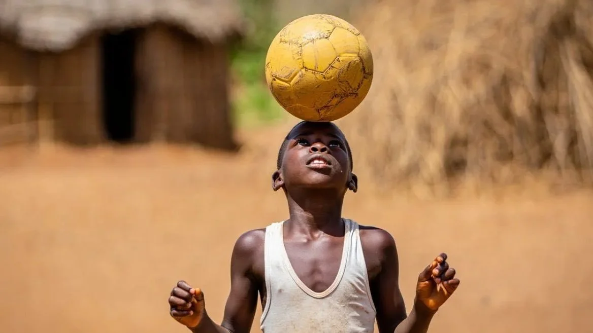 Young boy balancing a yellow soccer ball on his head outdoors.