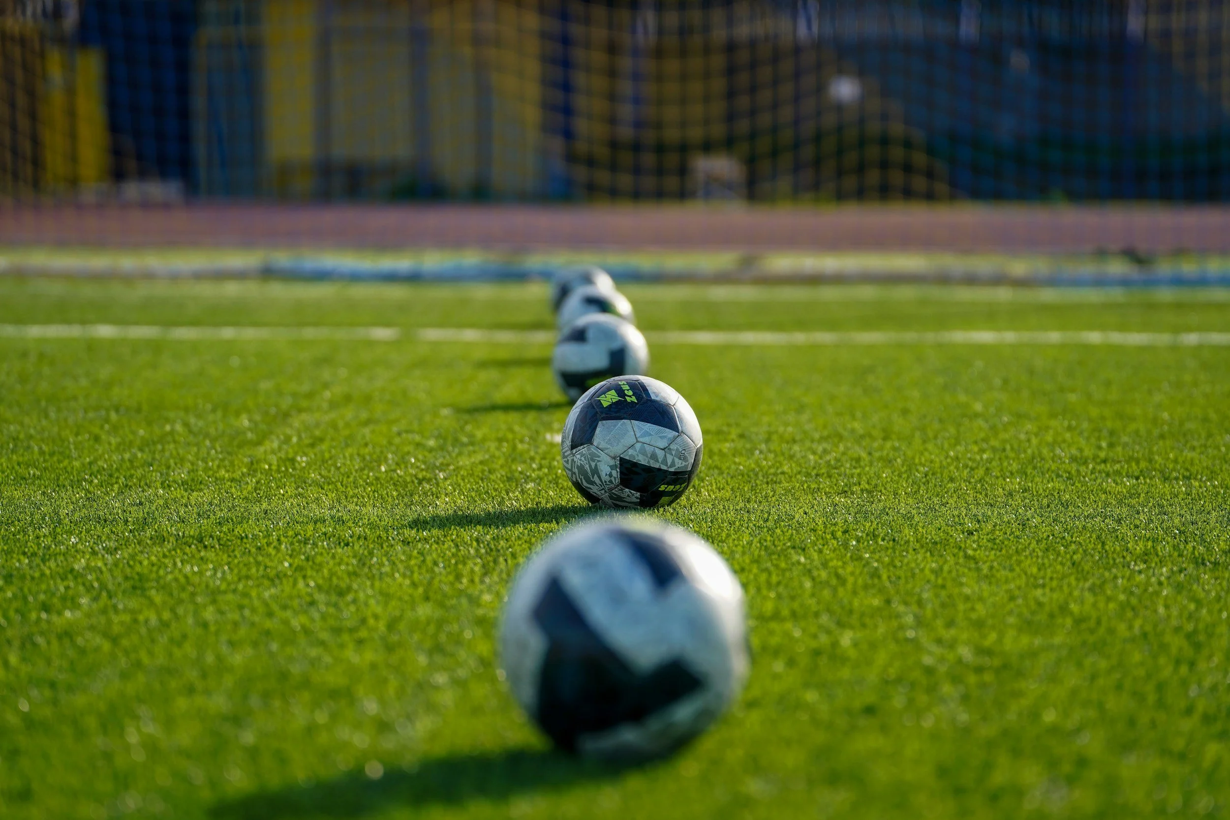Multiple soccer balls lined up on a green soccer field, with a goal net in the background.