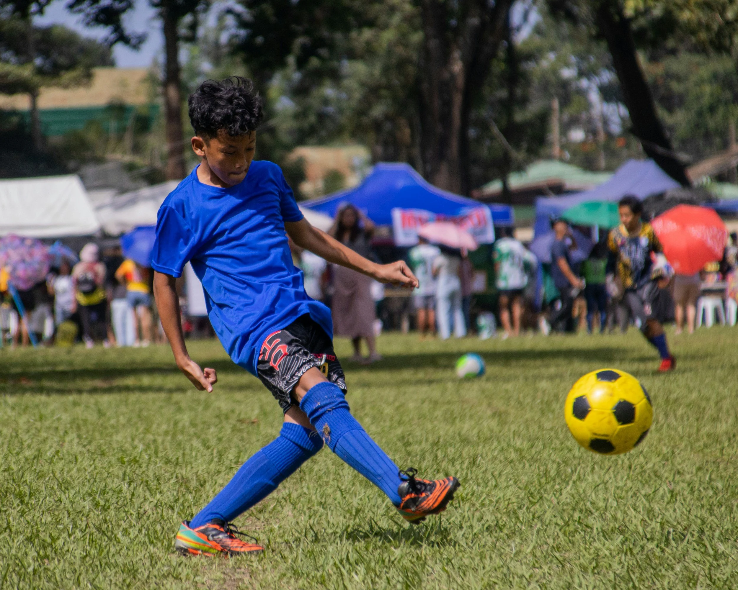 Child in blue sports uniform kicking a yellow and black soccer ball on a grassy field, with a crowd of people, tents, and trees in the background.
