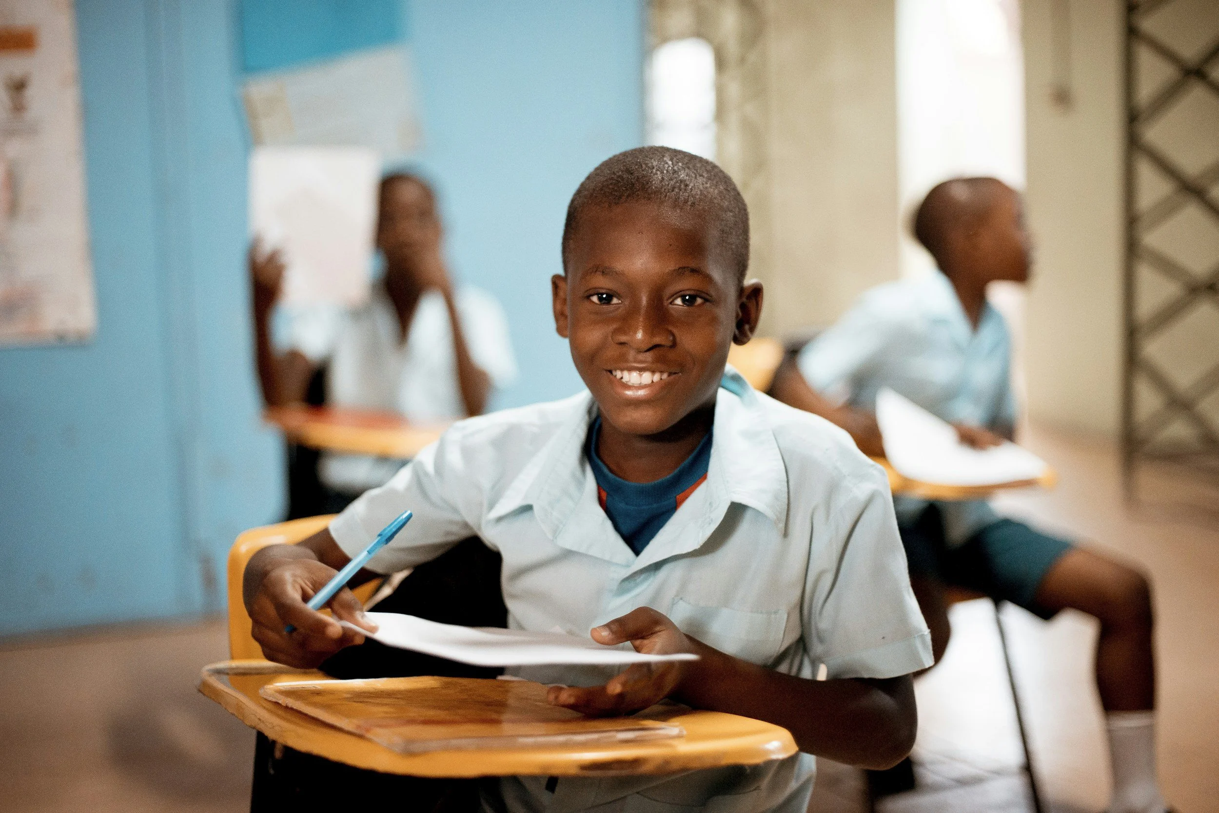 A young boy in a light blue school uniform sitting at a desk in a classroom, smiling at the camera with a pen and paper in front of him. Other students are visible in the background.