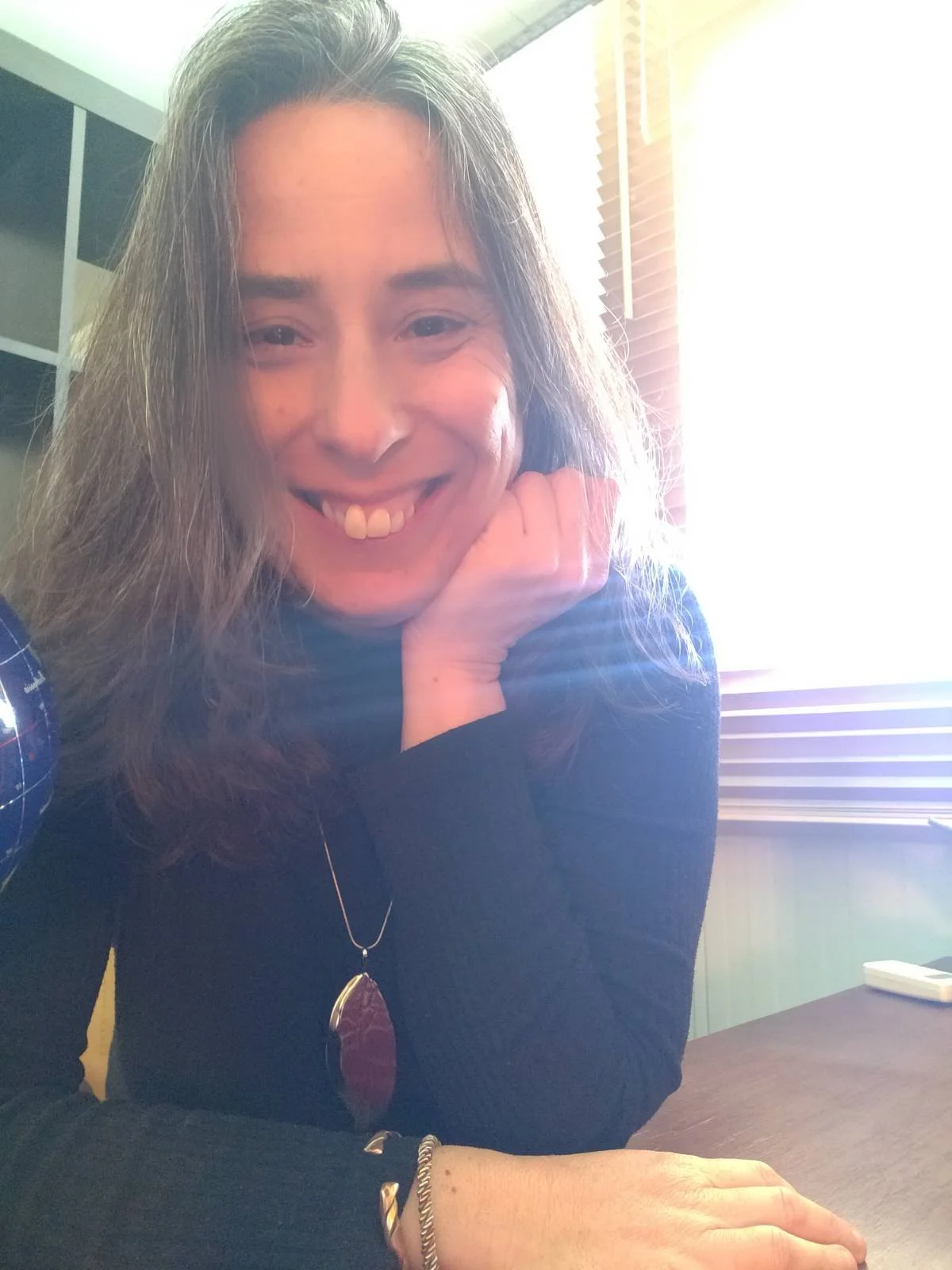 Close-up of Sandra Gonçalves , psychologist, resting her chin on her hand, sitting at a desk in front of a window with blinds.