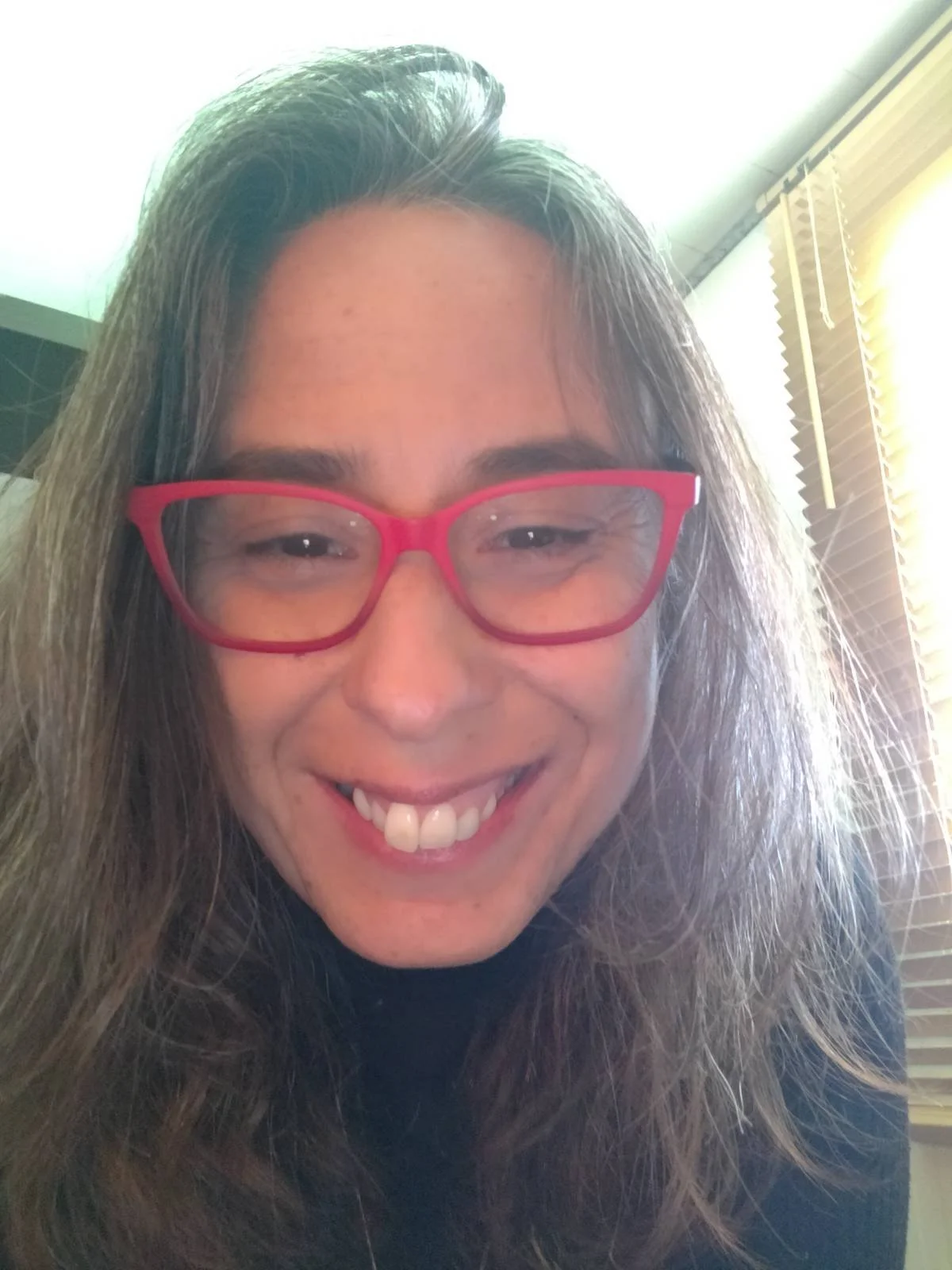 Close-up of Sandra Gonçalves, wearing red glasses, indoors near a window with wooden blinds.