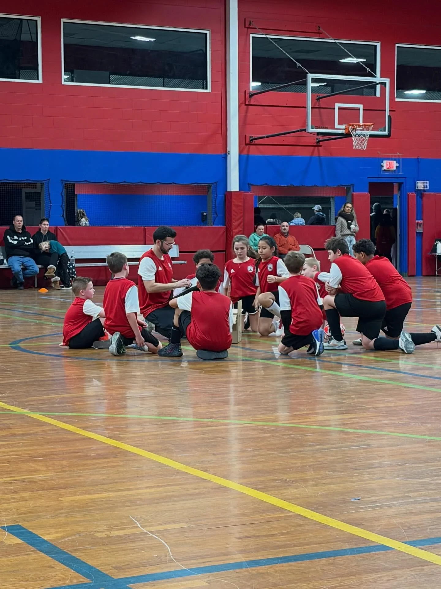 A group of children and a coach in a gymnasium, participating in a youth basketball practice or game, with some adults sitting and standing on the sidelines.