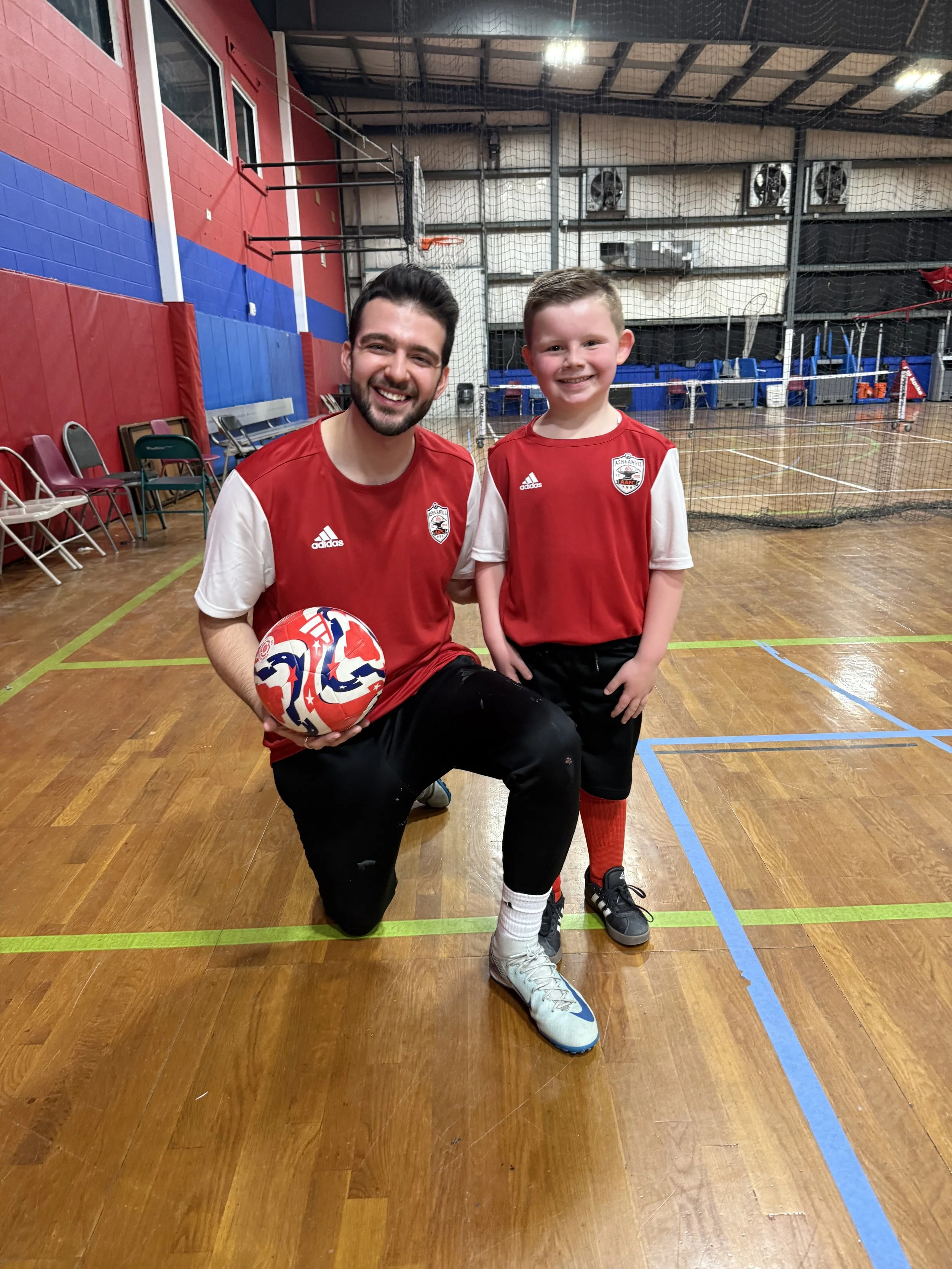 A man and a young boy in sports uniforms inside an indoor gymnasium, posing for a photo with a volleyball. The man is kneeling with a smile and the boy is standing next to him, also smiling.