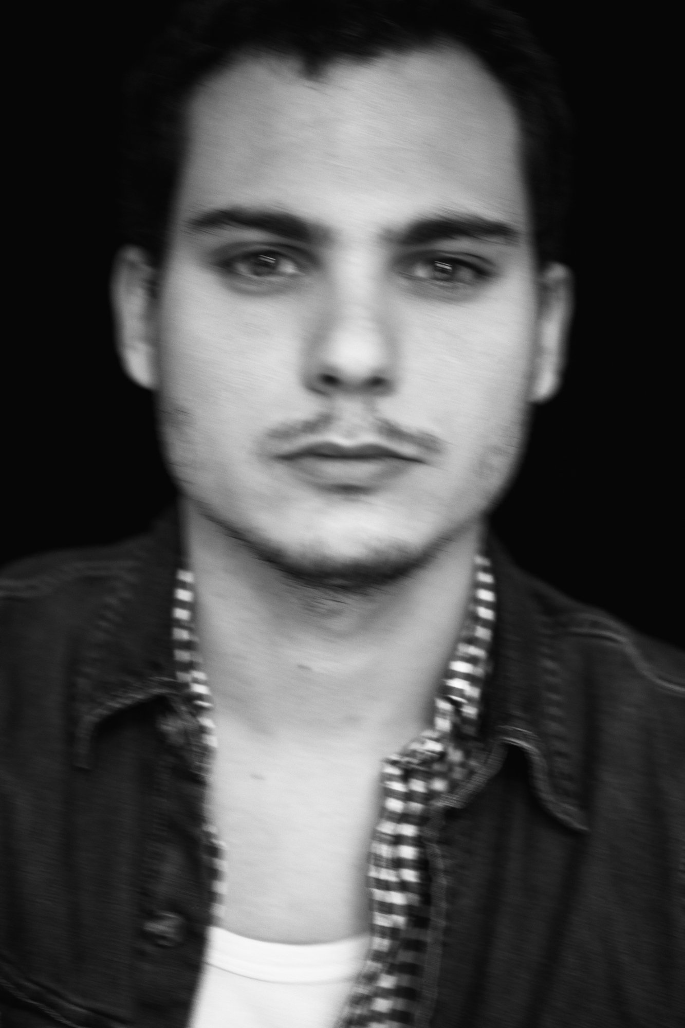 Portrait en noir et blanc d'un jeune homme avec des cheveux foncés, portant une chemise à carreaux et une veste en denim, regardant vers l'objectif, fond noir.