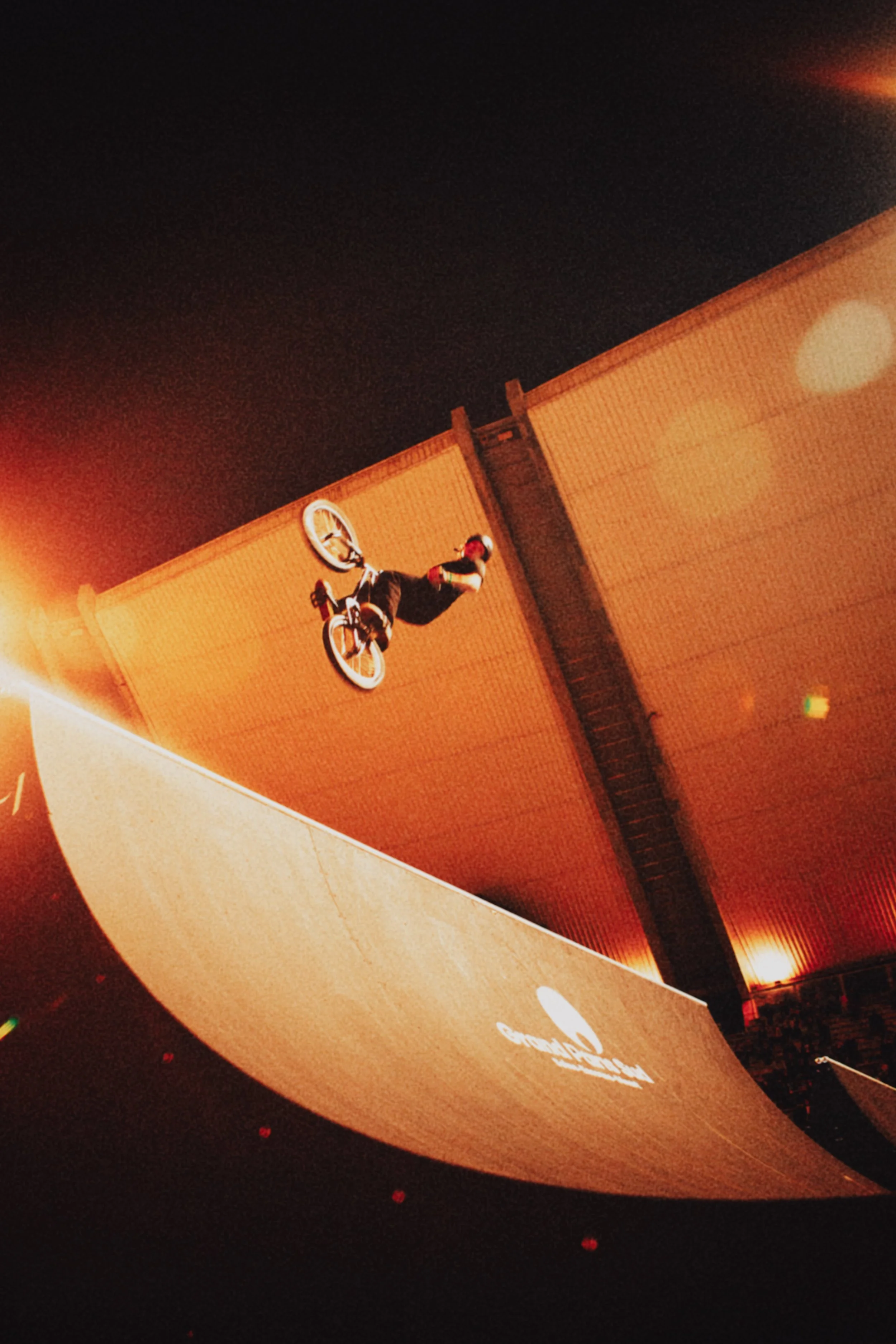 Un fille effectuant une figure de BMX dans un skatepark la nuit, avec un éclairage orange. La photo est prise de côté, montrant la fille en l'air avec le BMX.