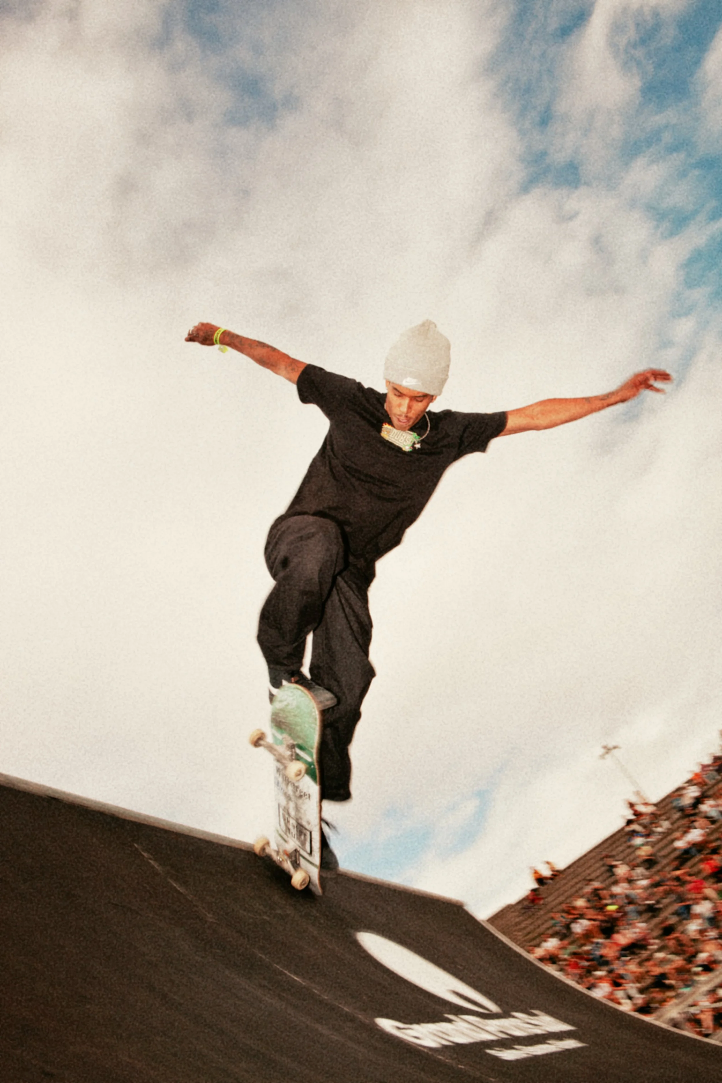 Un skateur effectue un saut en longueur sur une rampe dans un skatepark avec un ciel partiellement nuageux en arrière-plan et une foule regardant depuis en bas.