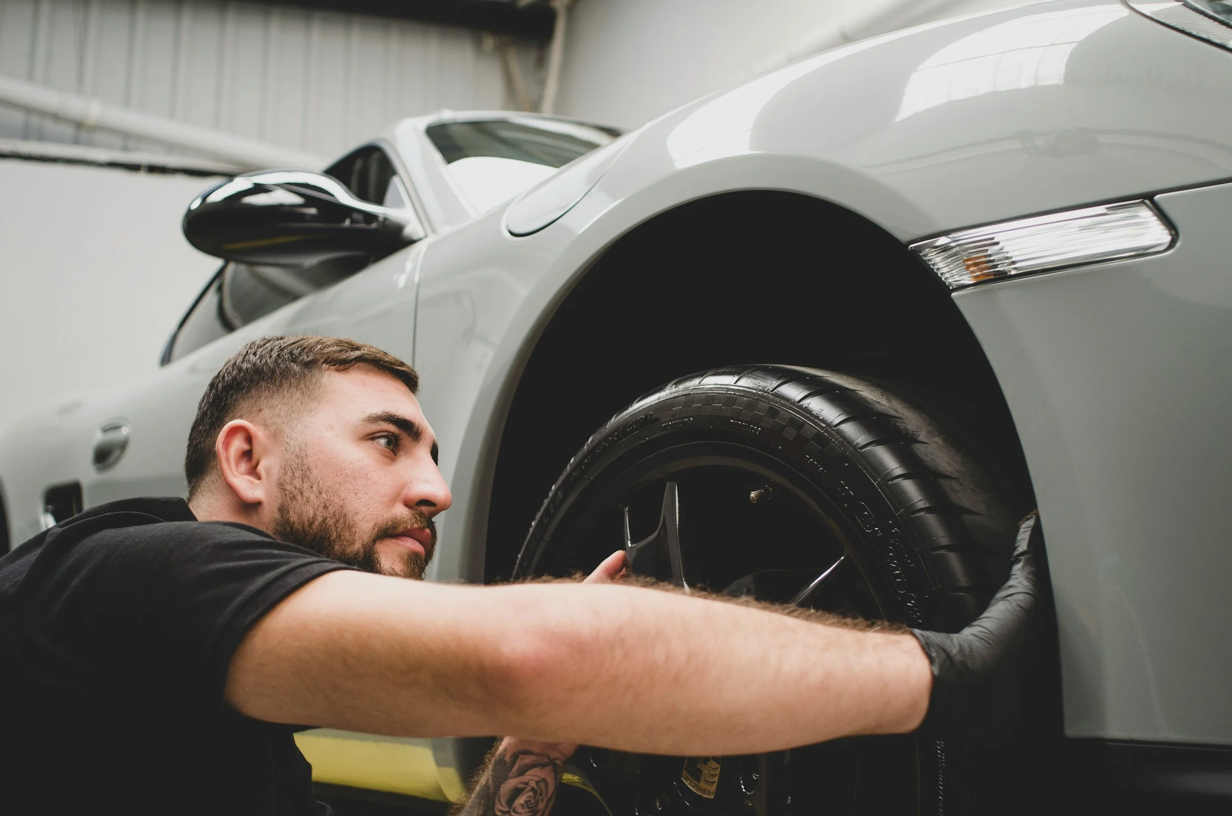 A man fixing or inspecting the wheel of a silver sports car in a garage or workshop.