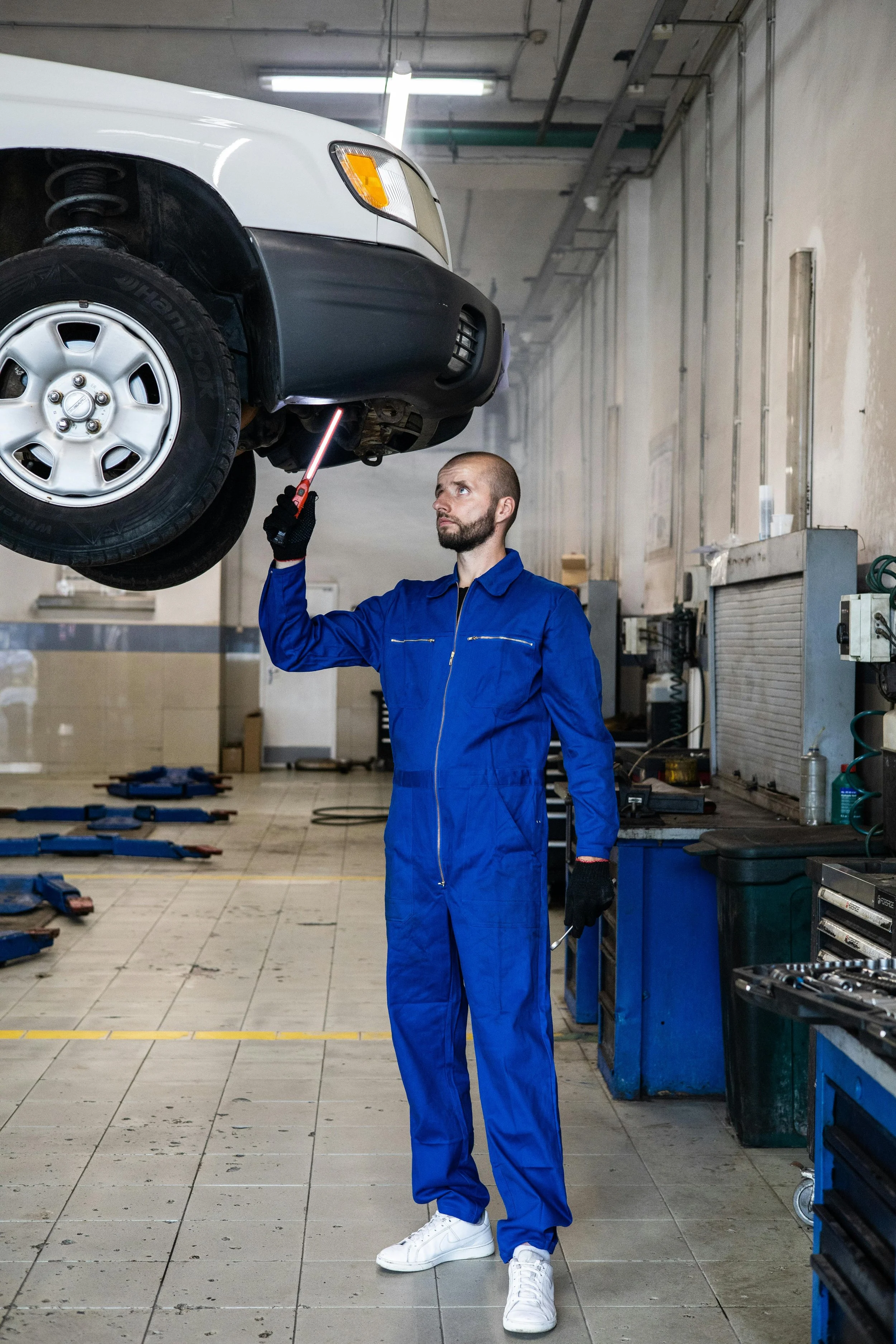 A mechanic in a blue jumpsuit and white sneakers inspects or repairs the underside of a white vehicle raised on a hydraulic lift in an auto repair shop.