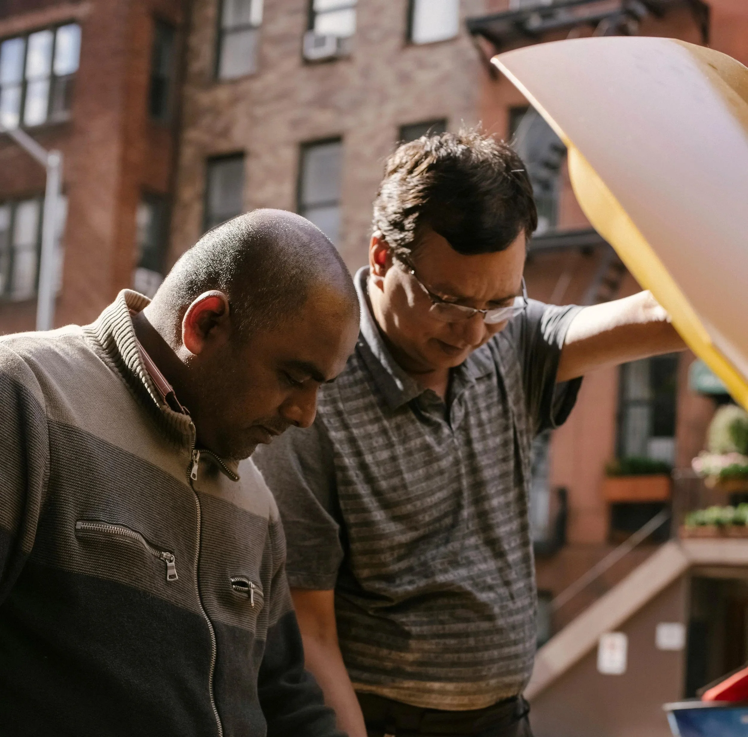 Two men are looking into the open hood of a car parked in an urban residential area with brick buildings in the background.