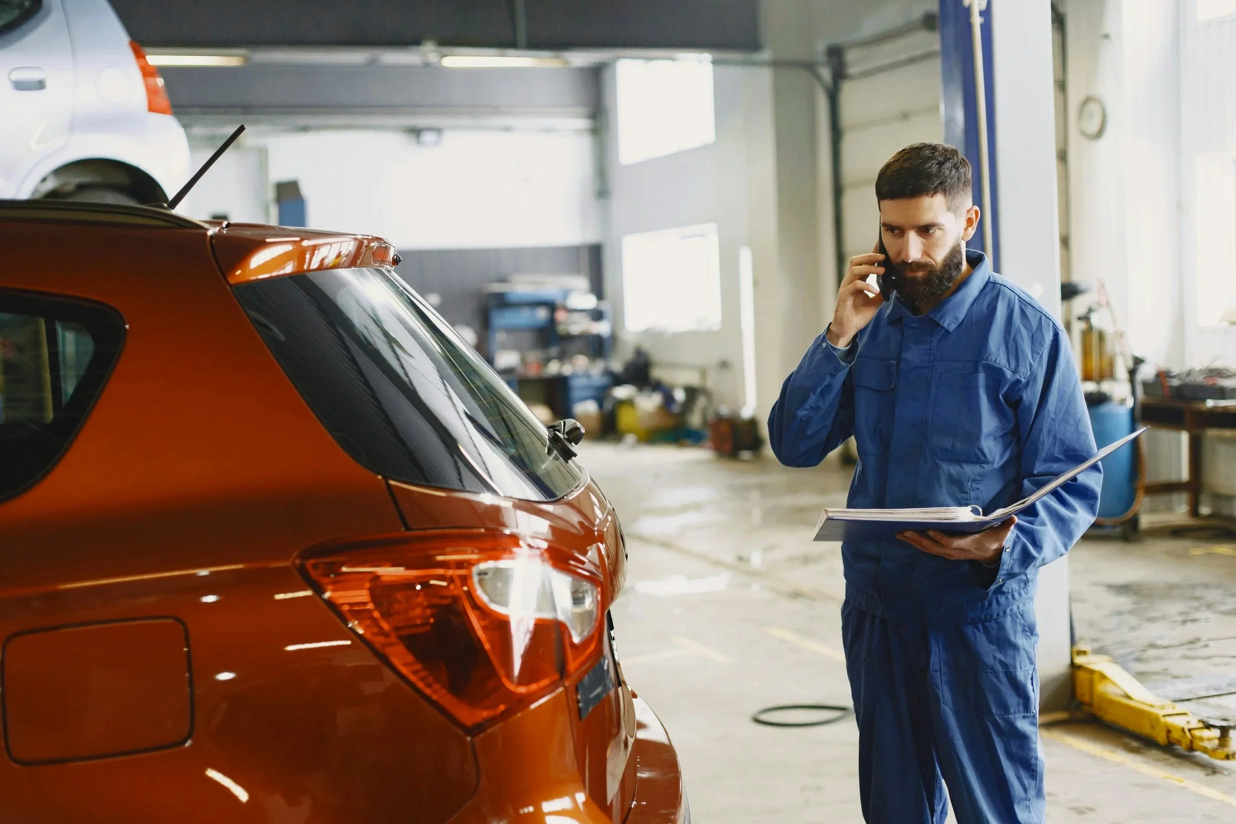 A man in blue overalls talking on the phone while holding a clipboard in an automotive repair shop, standing next to a brown car.