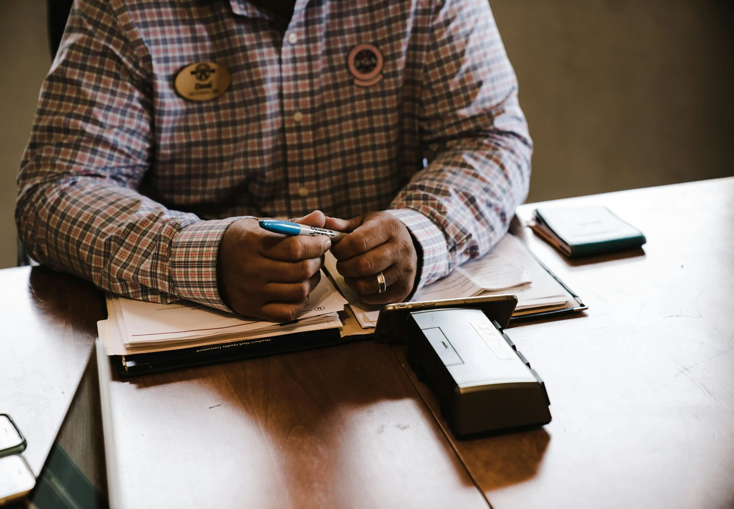 A person sitting at a wooden desk, wearing a plaid shirt with a name badge and a pin, holding a pen. There are papers, a clipboard, a smartphone, and a cardholder on the desk.