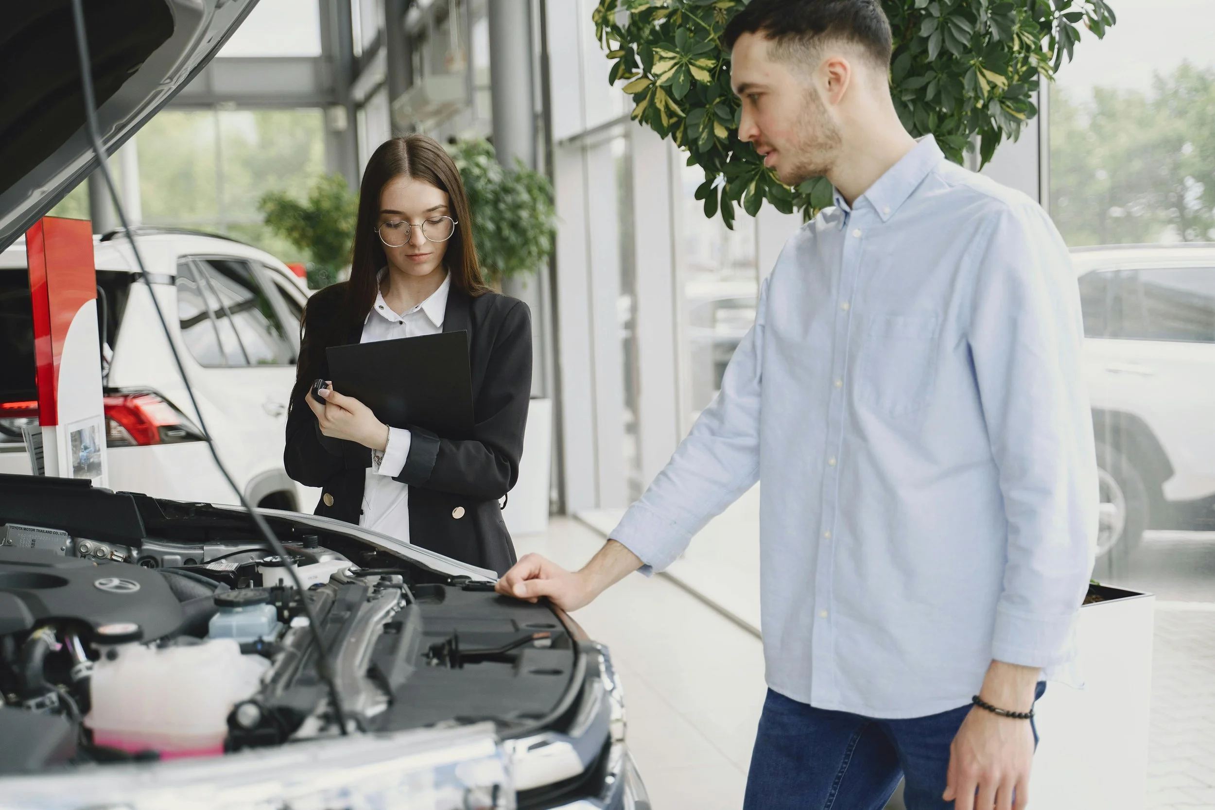 A man and a woman standing in a car showroom inspecting the engine of a car with the hood open. The woman is holding a clipboard and looking at the engine, while the man is gently touching the car's engine with his right hand.