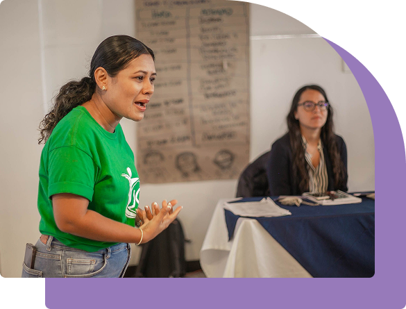 A woman in a green T-shirt speaking and gesturing during a group workshop, with another participant seated at a table in the background, demonstrating communication, collaboration and shared learning.