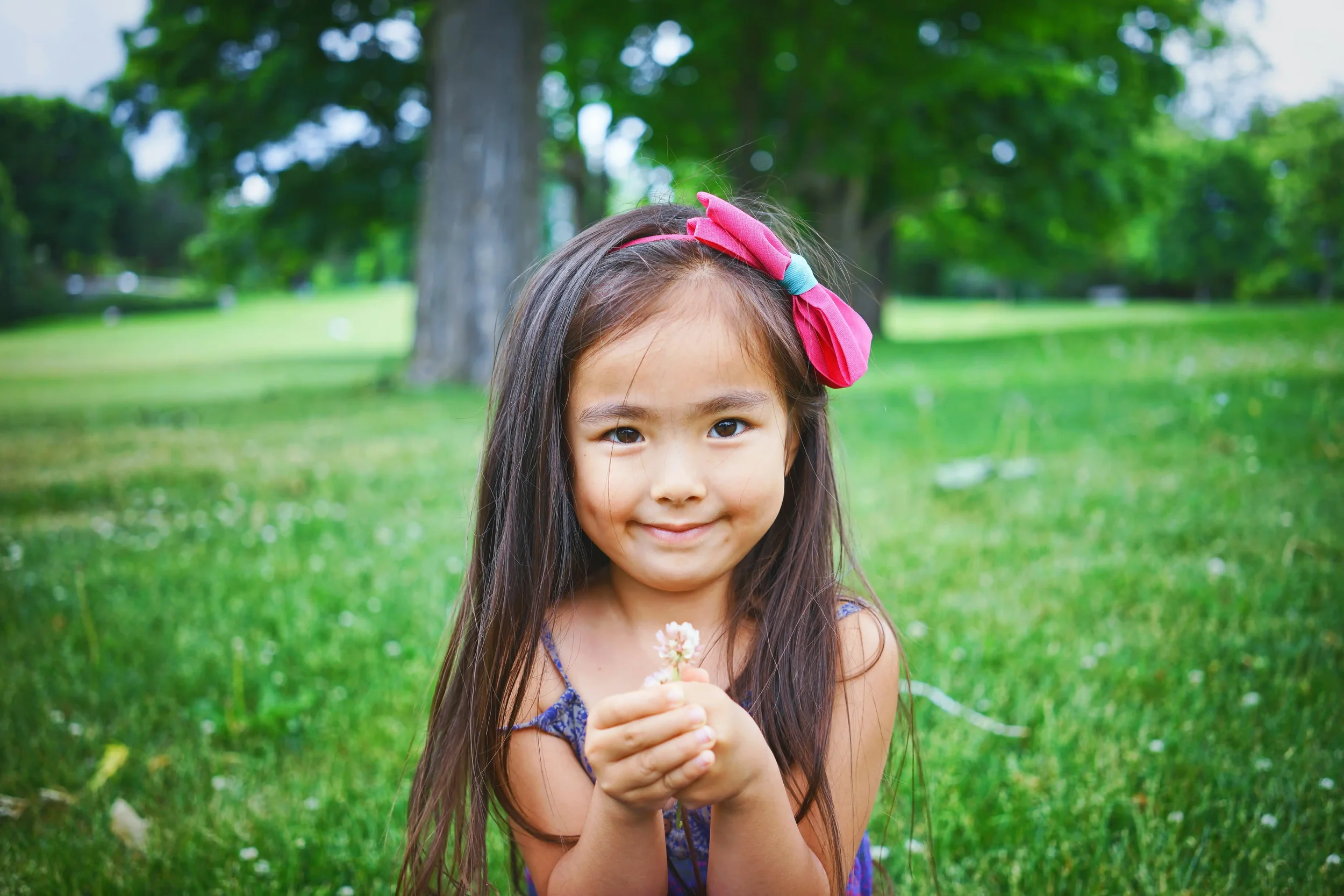 A young girl holding a small flower while sitting on the grass in a park.