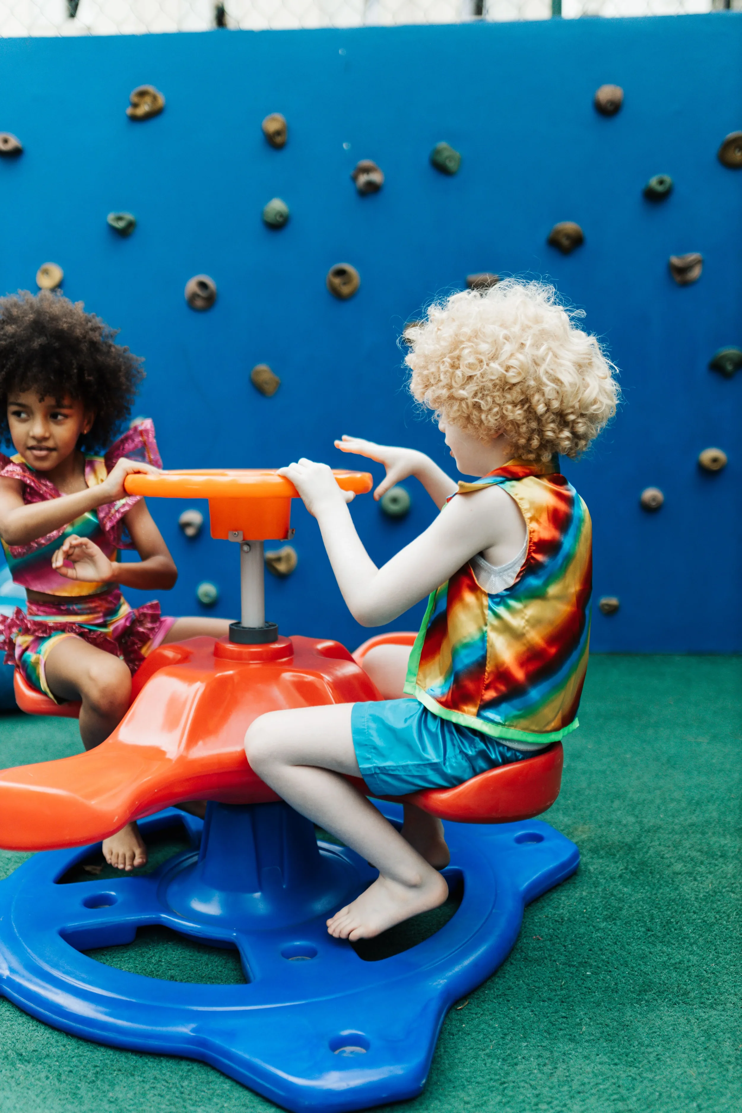 Two children playing together on a colourful playground seesaw in front of a climbing wall, representing childhood development, inclusion and the importance of play.