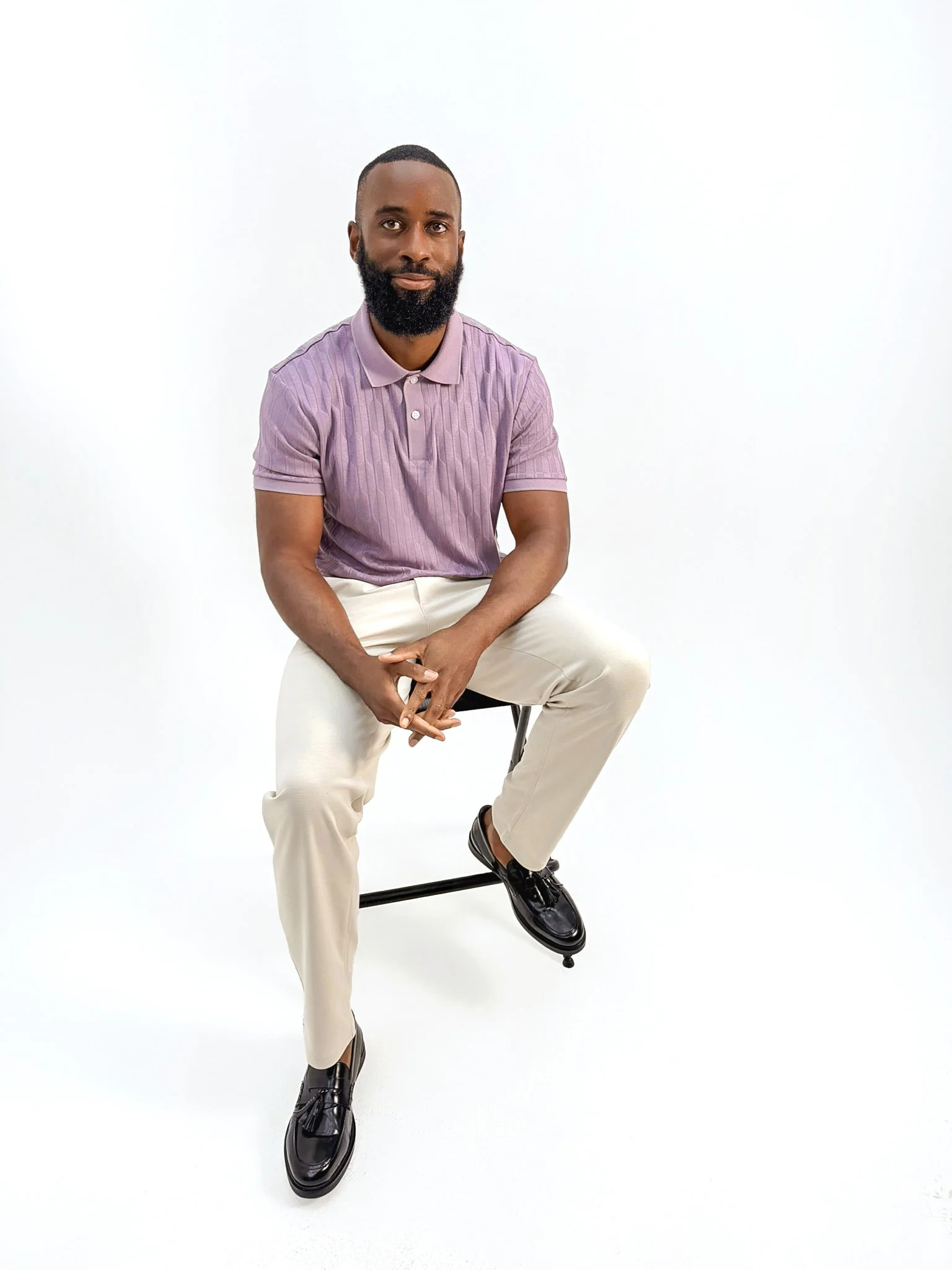 Professional portrait of 4REAL's CEO seated on a stool, wearing a lilac polo shirt and cream trousers against a clean white background.