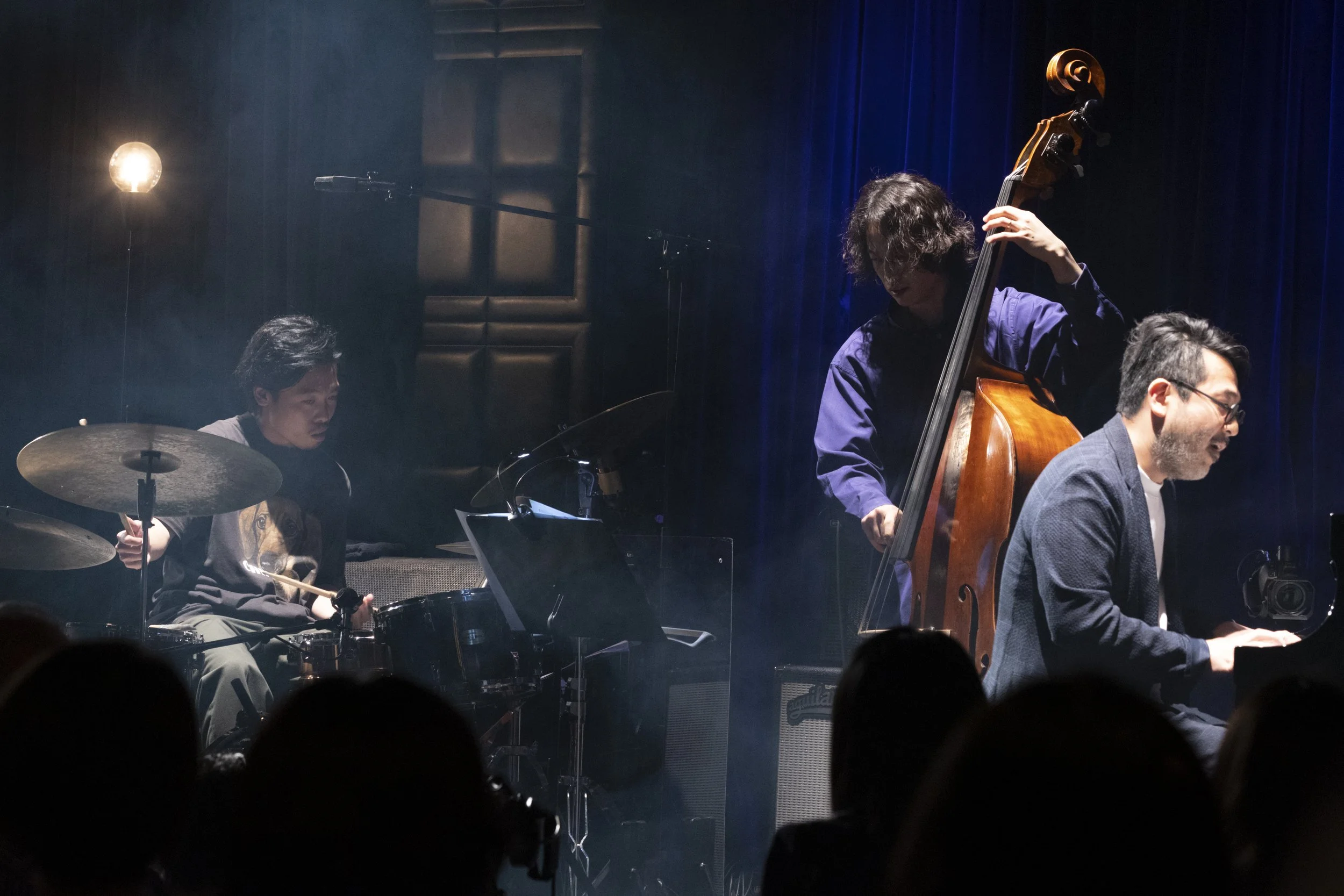 Three musicians performing on stage: a drummer Shun Ishiwaka, a double bass player Takashi Sugawa, and a pianist Masaki Hayashi, with blue stage curtains in the background.