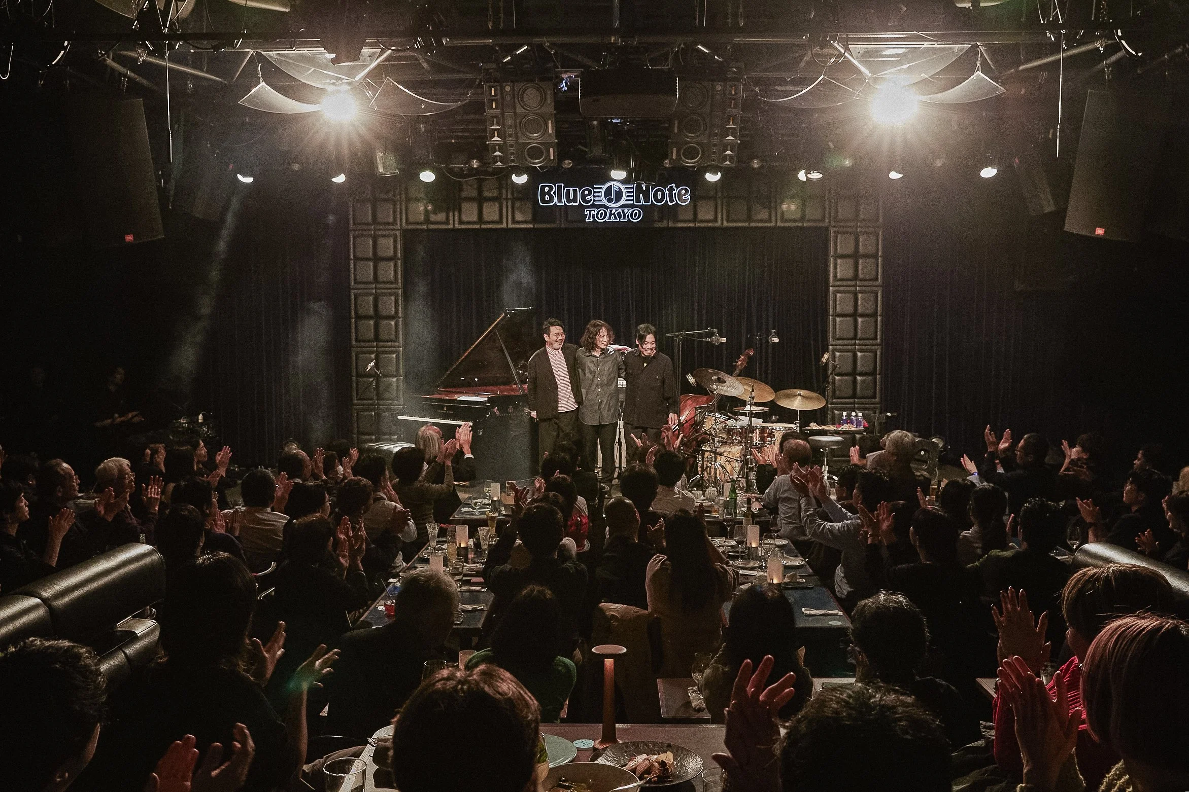 A live music performance at Blue Note Tokyo with three musicians on stage, audience clapping, stage lights shining, and a grand piano and drums visible on stage.