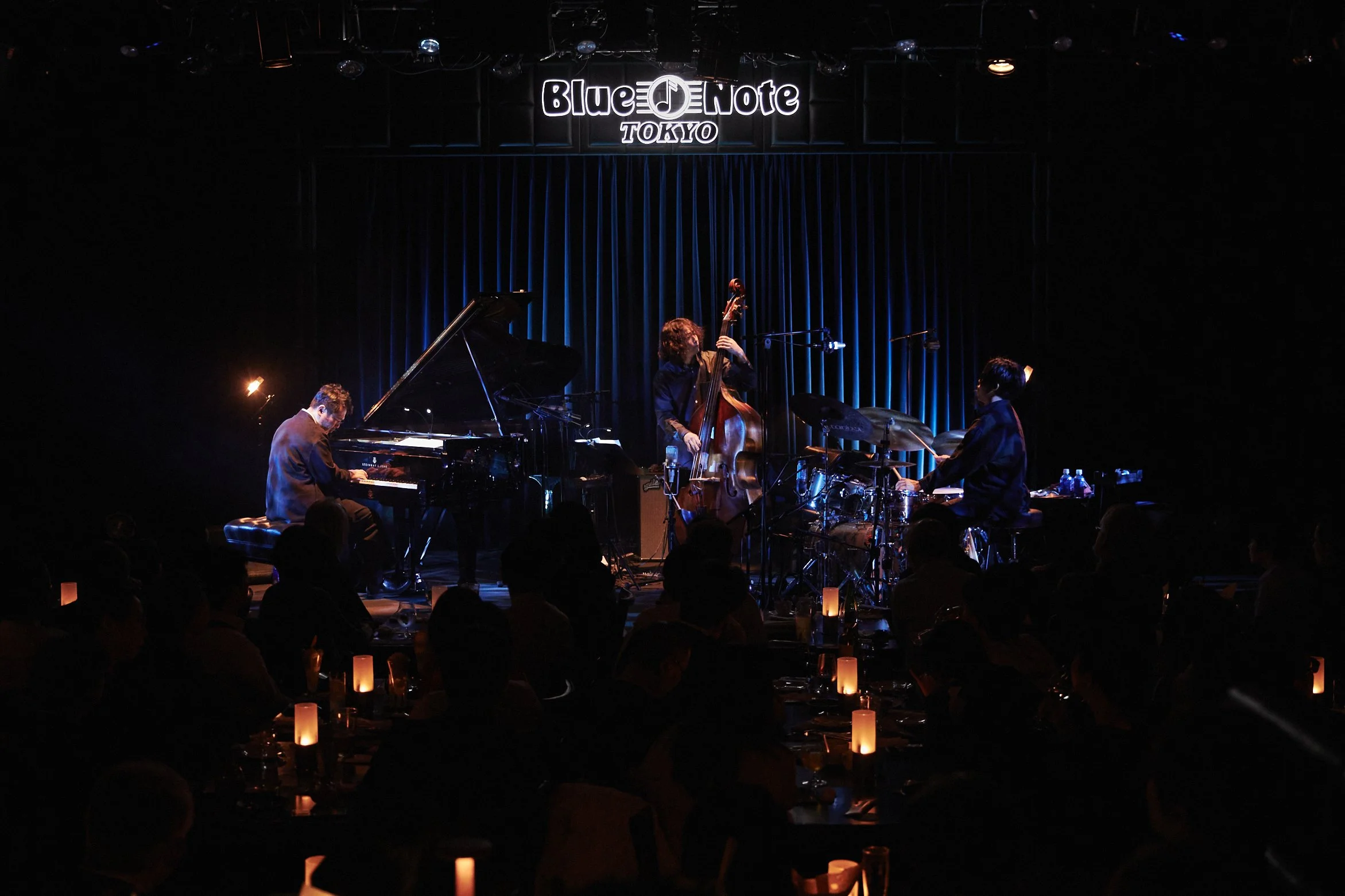 Jazz band performing on stage at Blue Note Tokyo with a pianist, double bassist, and drummer, audience seated in front, dim lighting, candles on tables.