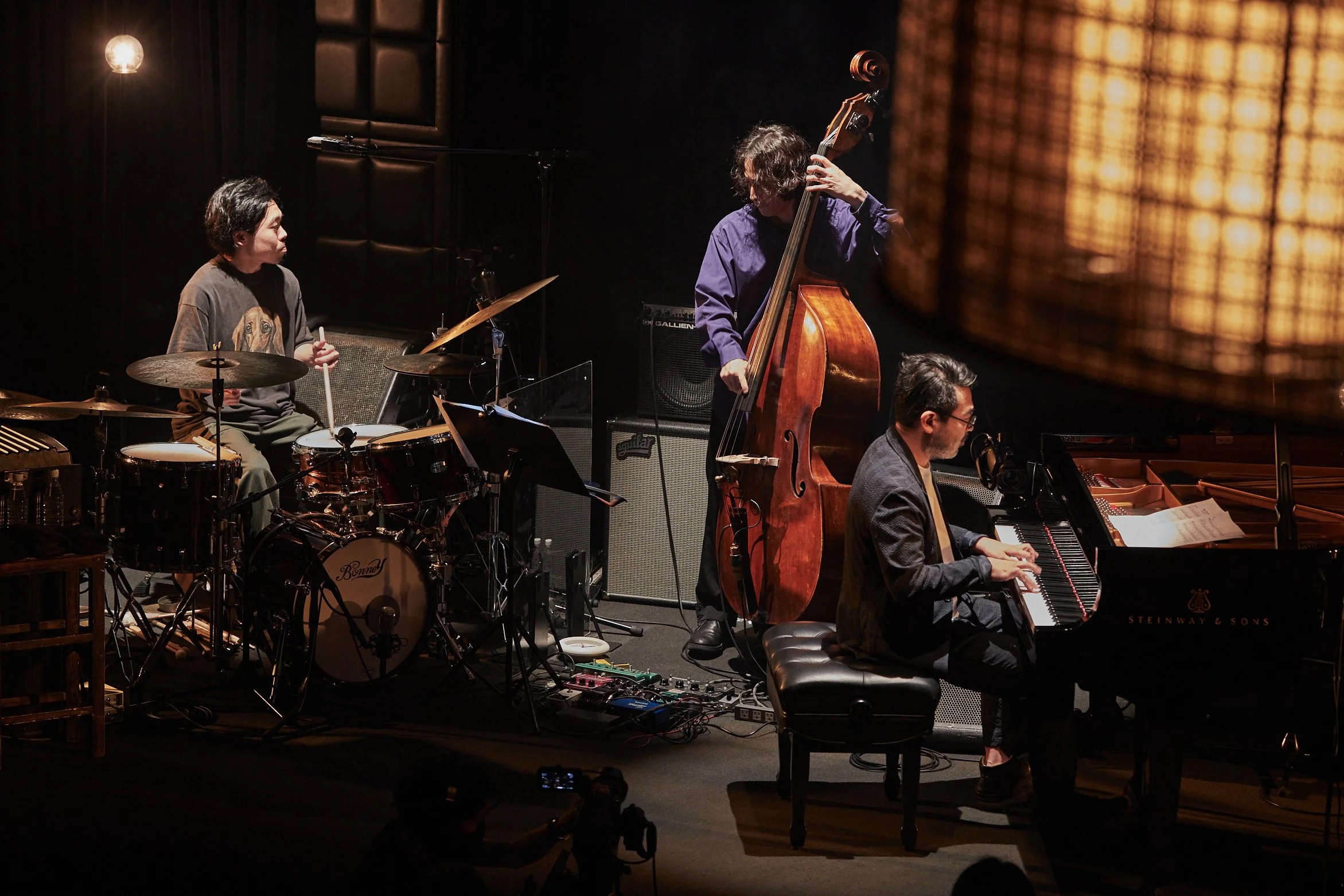 A jazz band performing on stage with three musicians: a drummer, a double bass player, and a pianist, under warm stage lighting.