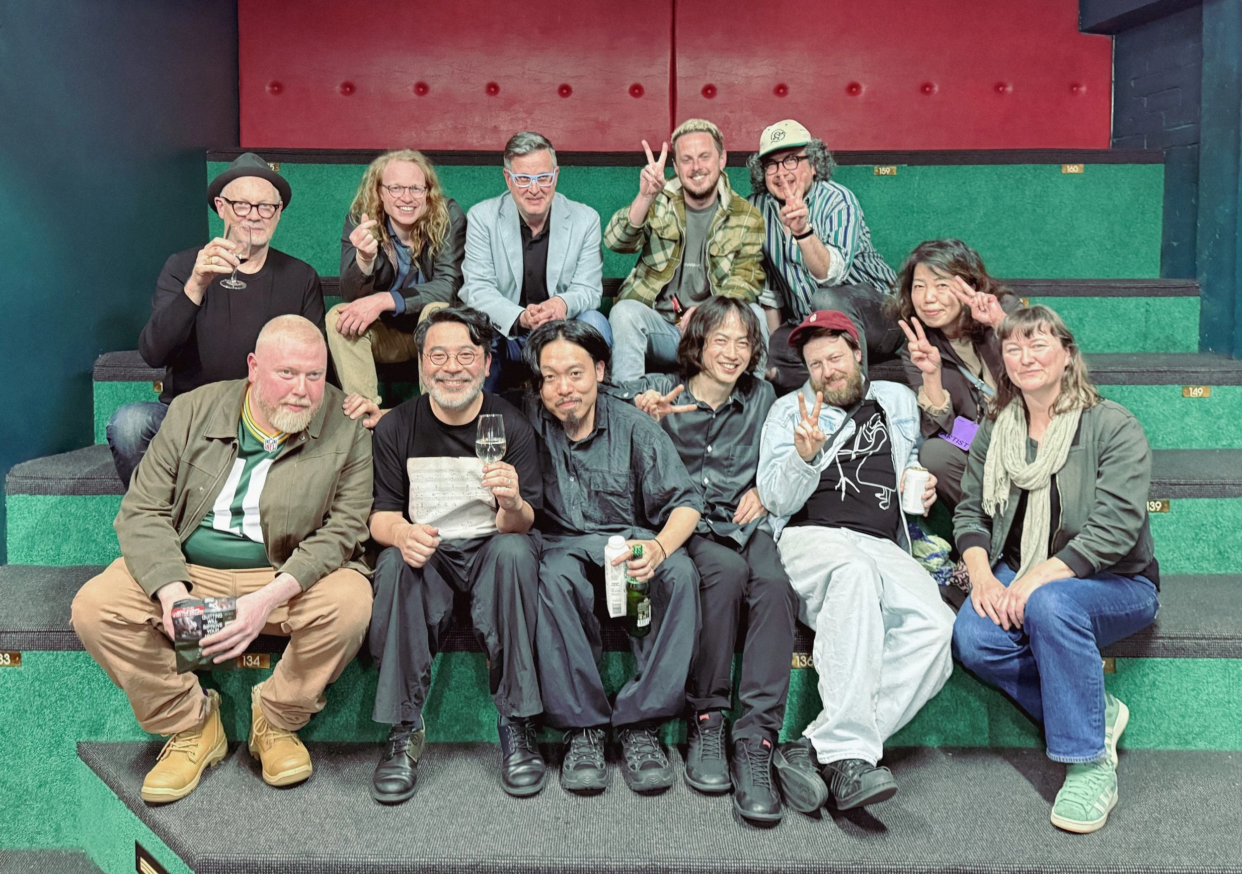 A group of 13 musicians from Melbourne and Tokyo sitting on green tiered theater seats, some holding drinks, smiling, and making peace signs in a casual indoor setting.