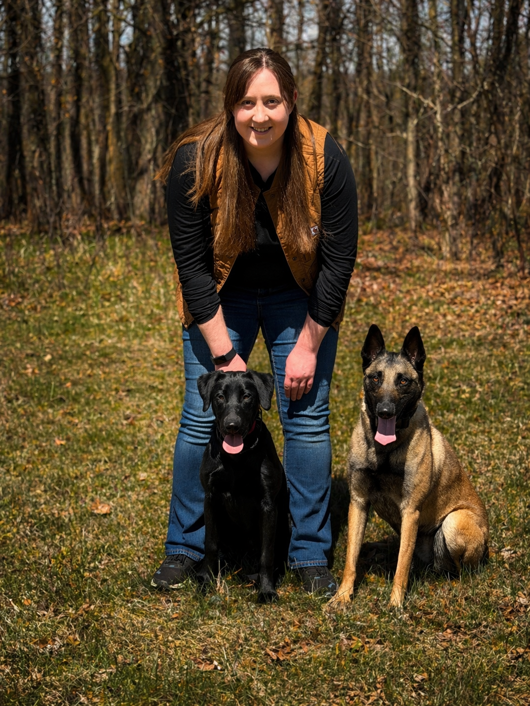 A woman with long, brown hair wearing a black shirt and brown vest, smiling, kneeling outdoors on grass with two dogs; one black Labrador with tongue out on the left, and one tan and with tongue out on the right, in a wooded area with trees in the background.