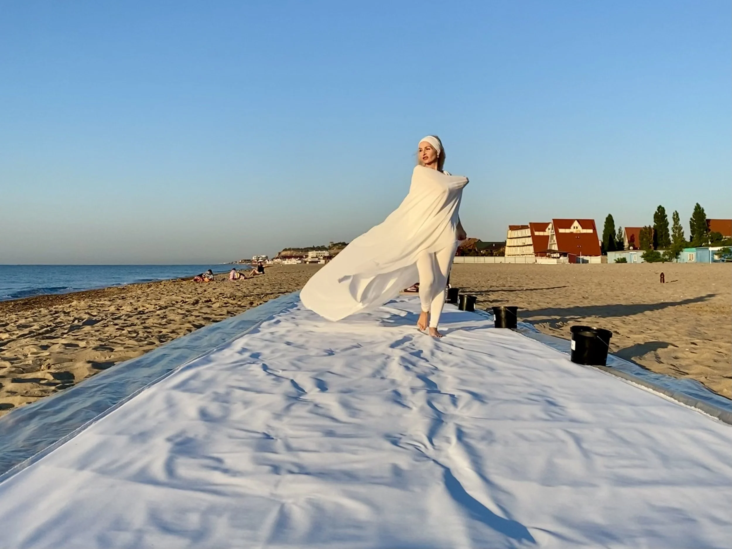 Iryna Sushelnytska in white clothing beside the continuous fifteen-metre canvas on the beach during the Nine performance.