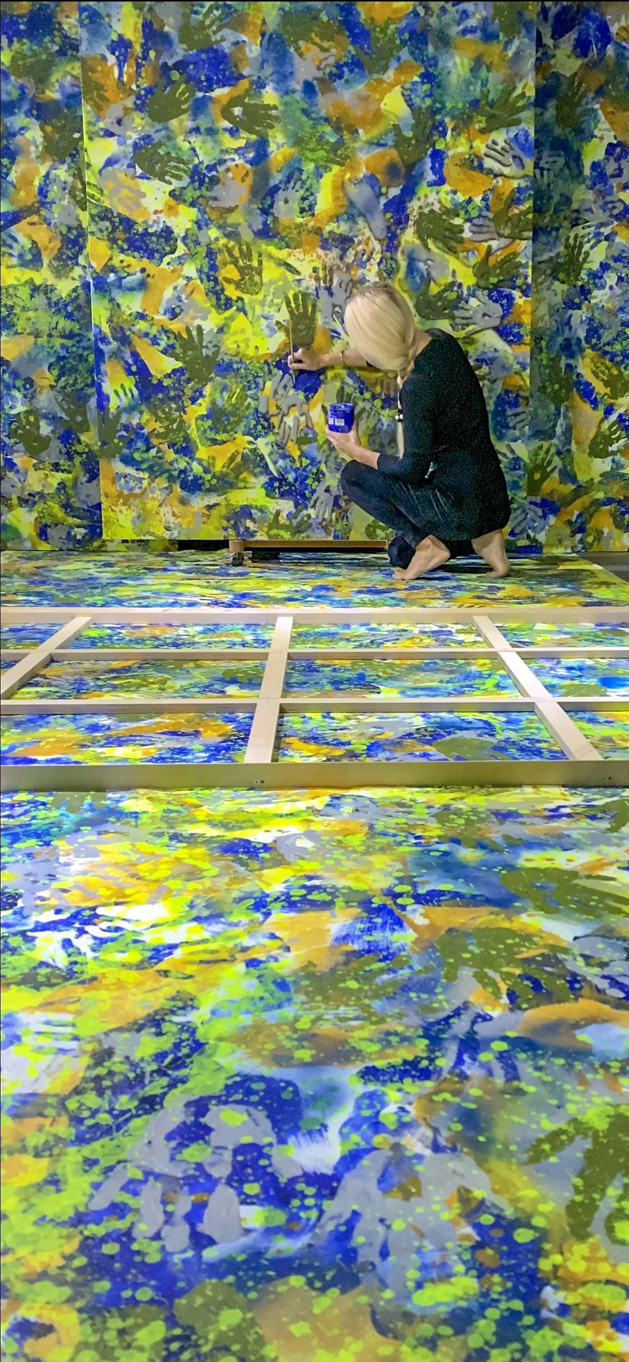 Iryna Sushelnytska working in the studio on Nine, with painted canvas sections on the floor and a large painted surface behind her.
