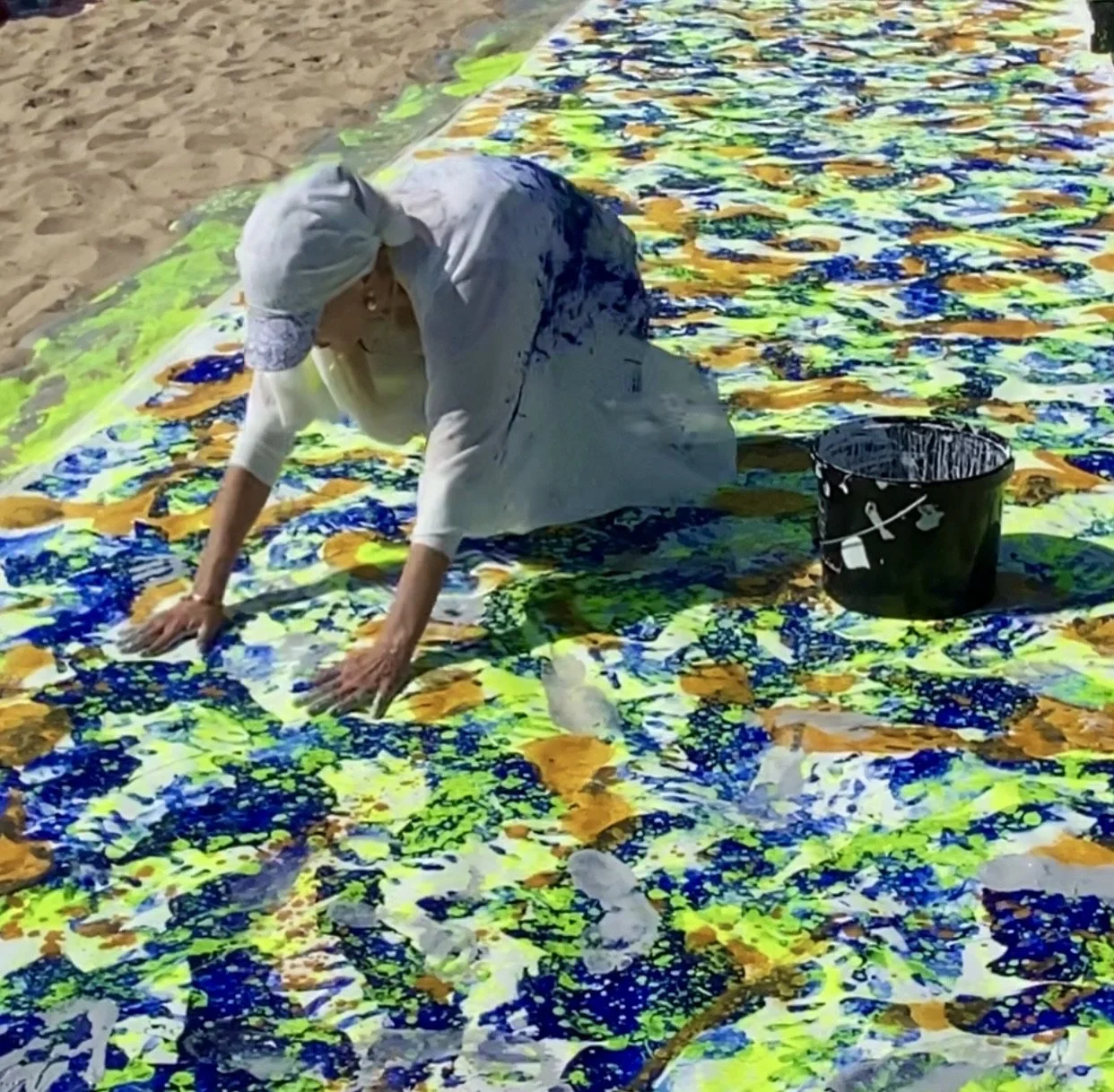 Iryna Sushelnytska kneeling on the painted canvas of Nine and pressing both hands into its blue, yellow and green surface.