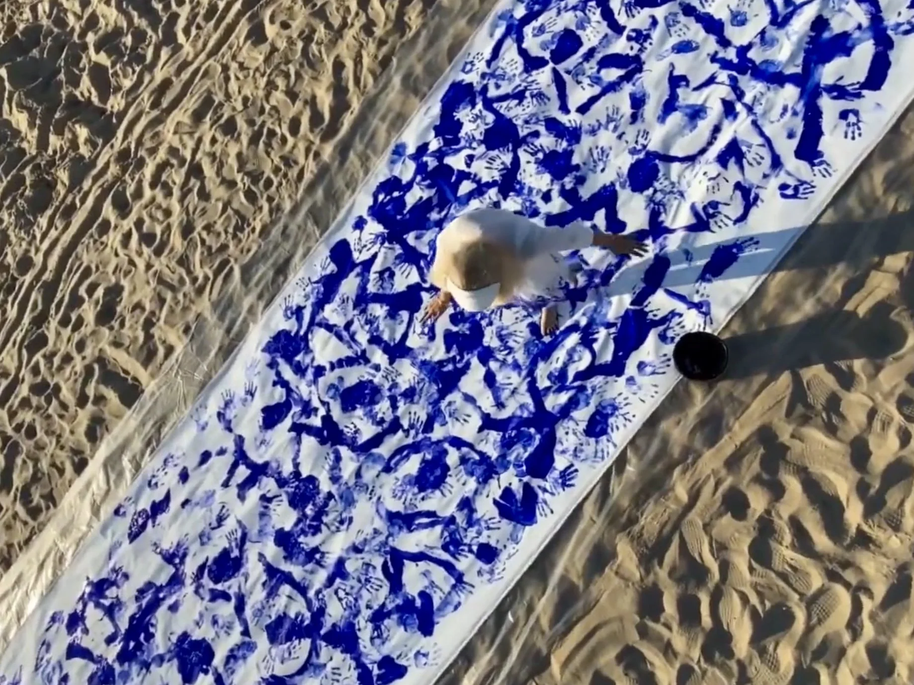 Overhead view of Iryna Sushelnytska working on the long white canvas of Nine on the sand, with dense blue marks across its surface.
