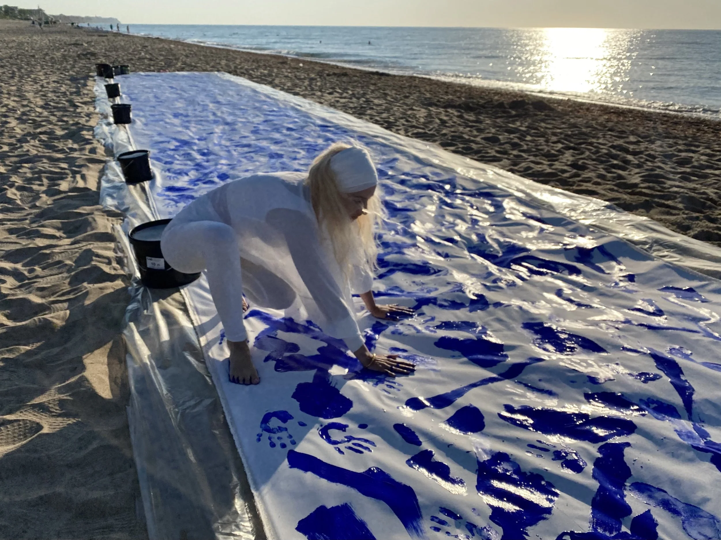 Iryna Sushelnytska crouching over the continuous canvas of Nine beside the sea, surrounded by blue marks on the white surface.