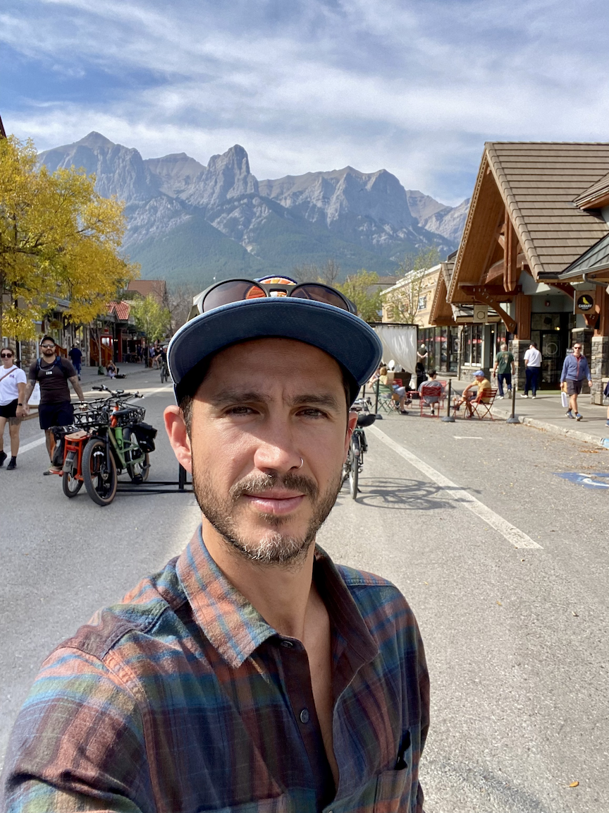 Selfie of a man with a beard wearing a plaid shirt, sunglasses on cap, and piercings, in a town with mountainous background, shops, and people walking.