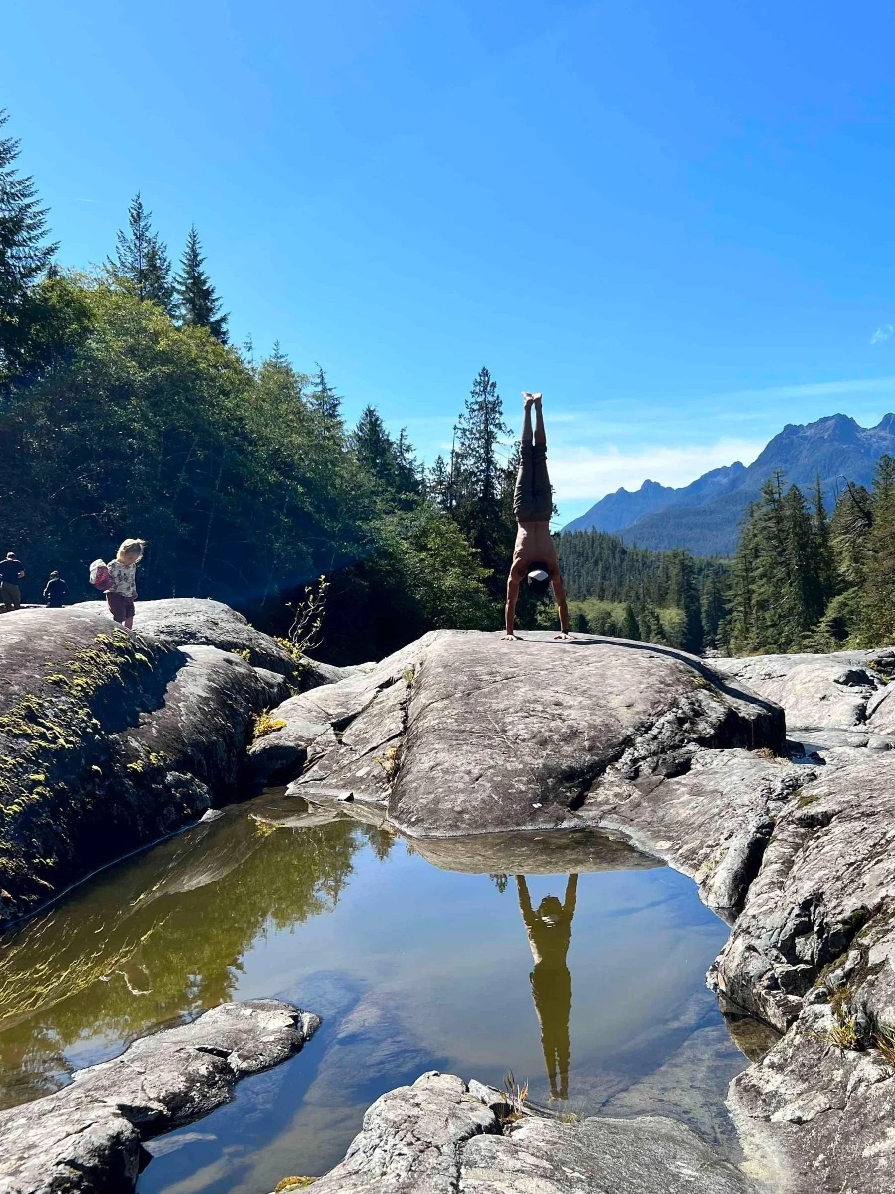 A man doing a handstand on a large rock by a river with mountains and trees in the background